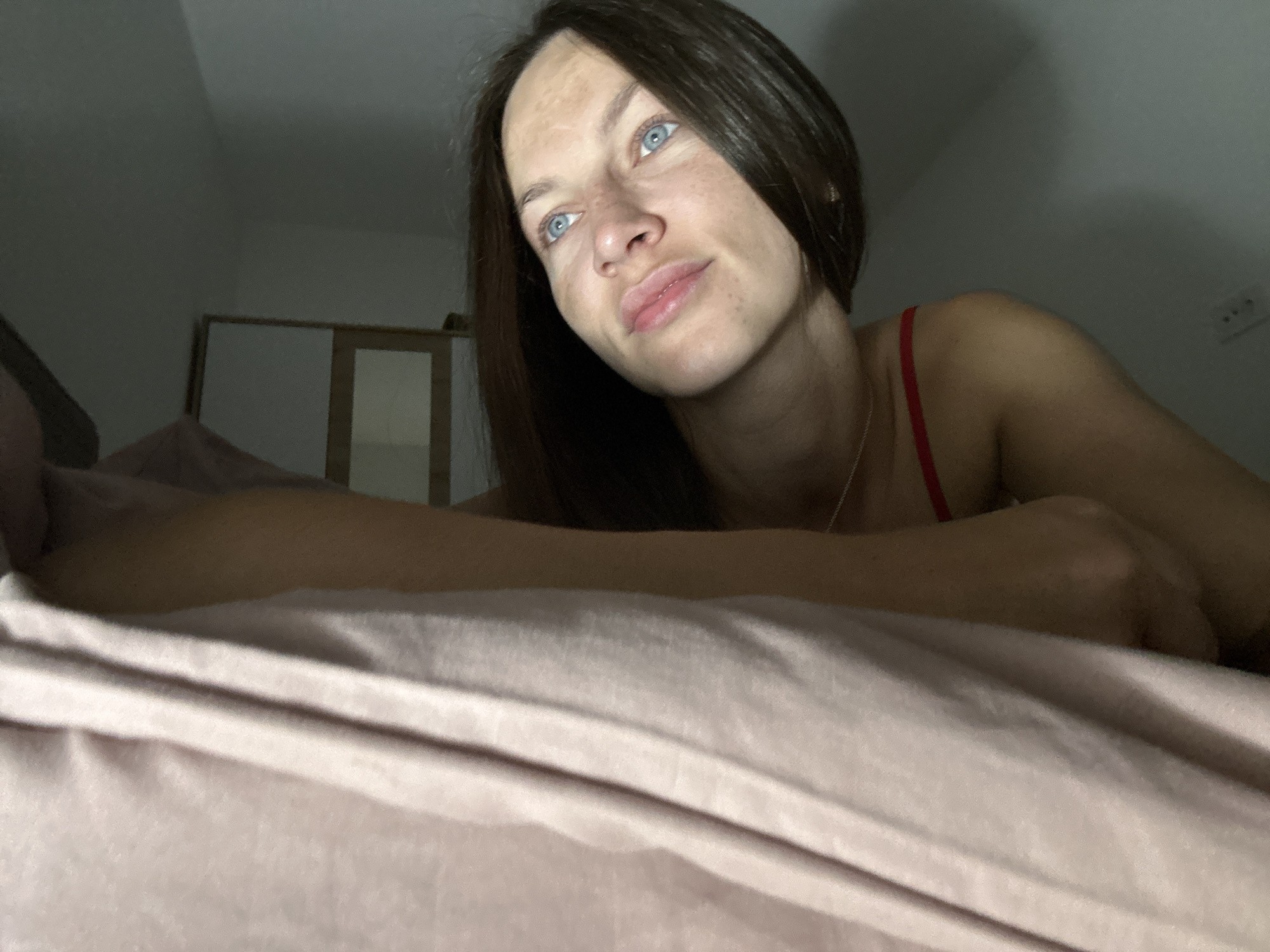 A woman with brown hair and blue eyes is lying in bed, looking up and to the side. She is wearing a red top and a silver necklace. The room is dimly lit, and the bed has pink sheets. In the background, there is a closet with white doors and a mirror. The scene is a bedroom in a home in Donji Pobori, Montenegro.