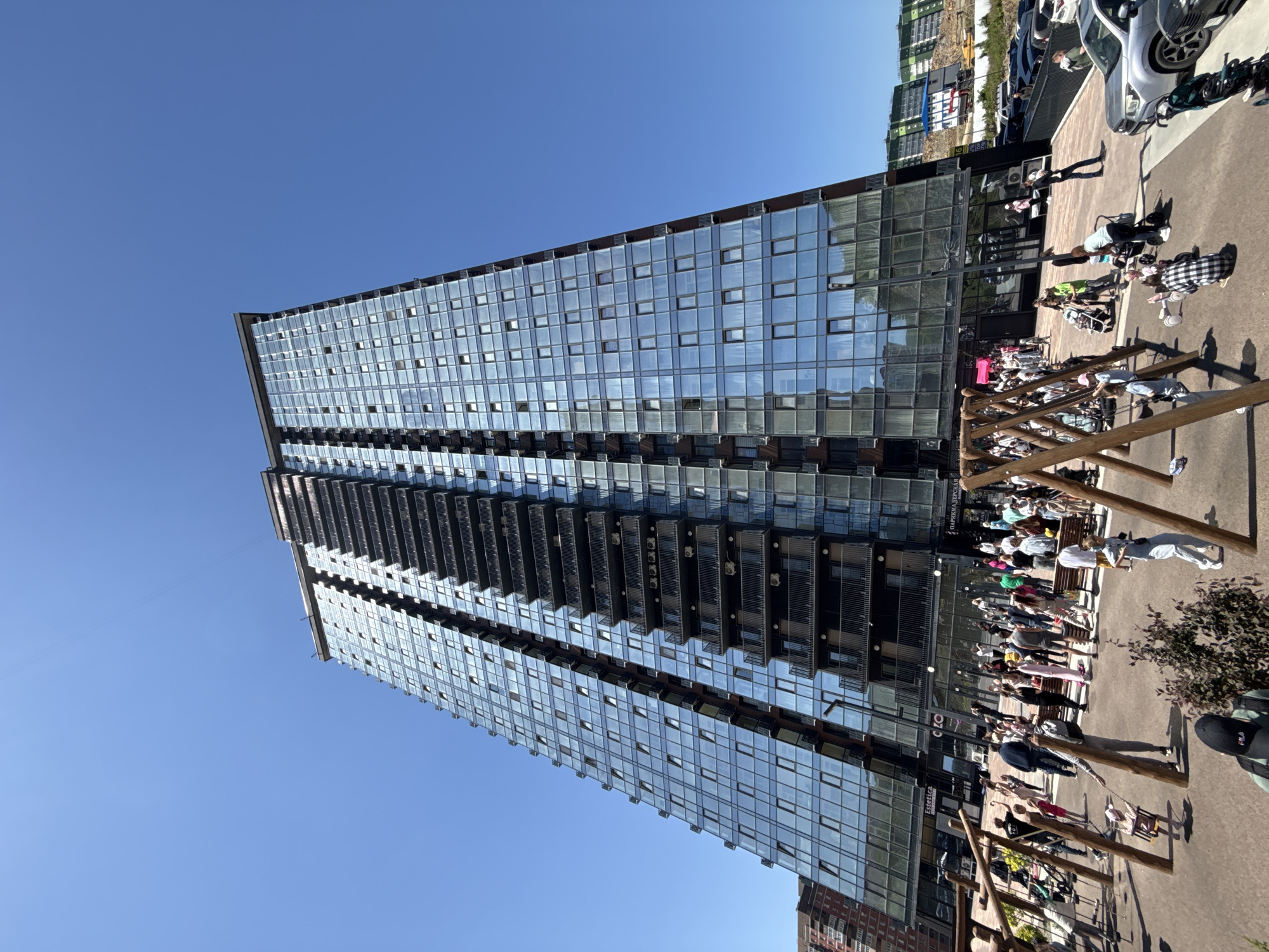 The image shows a tall, modern glass-facade building in Vladivostok, Russia. The building appears to be multi-story, with a primarily glass exterior featuring repeating rectangular windows. A crowd of people is visible in the foreground, gathered around a playground area with wooden swing sets. The scene suggests an urban environment, possibly a park or plaza adjacent to the building. A variety of cars are parked in the background, near another apartment building. The sky is clear and blue, indicating a sunny day. People are wearing casual clothing and appear to be standing and waiting. The photograph depicts a mixture of urban life and recreational space in Vladivostok.