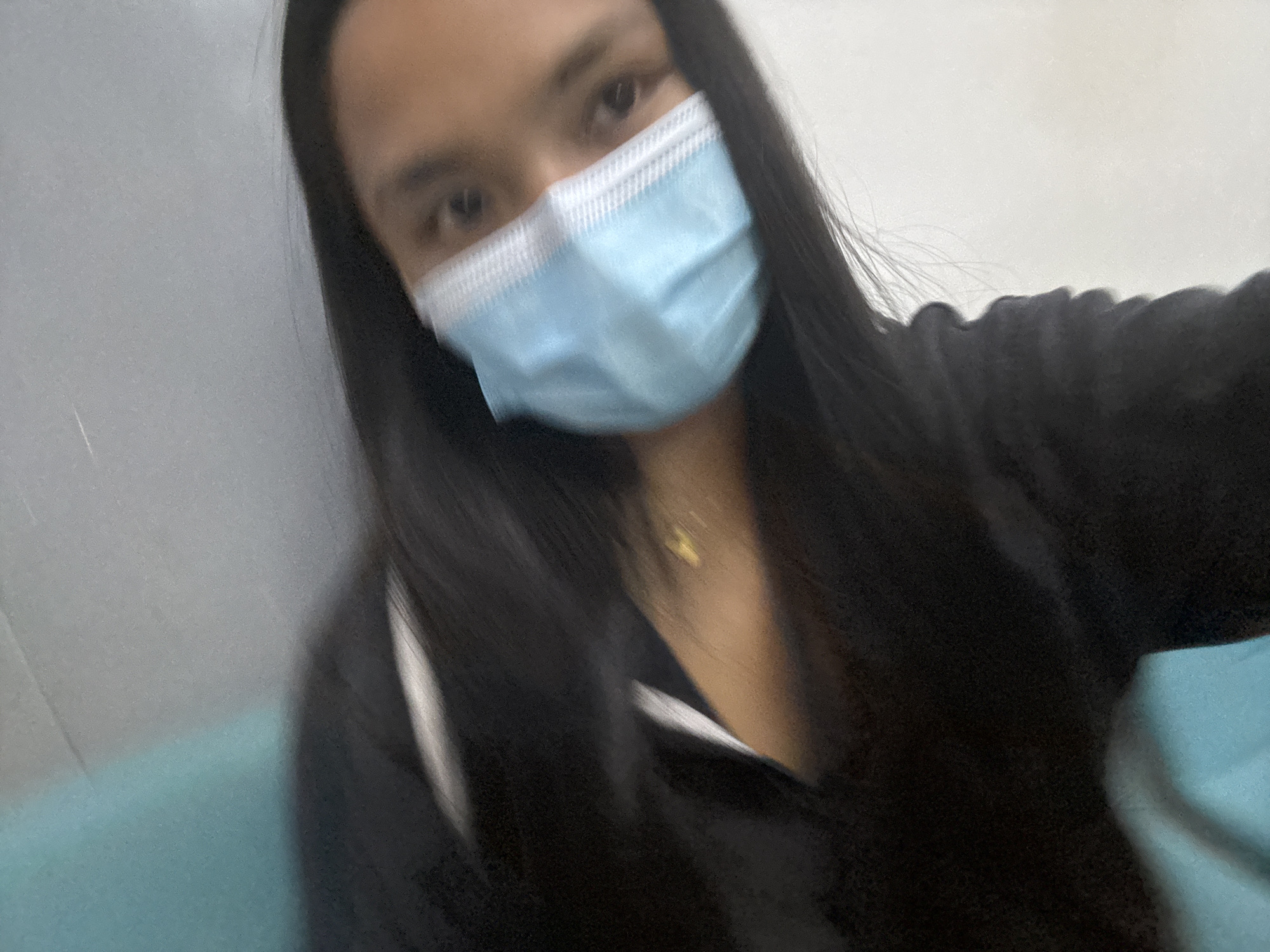 A person wearing a light blue surgical face mask and a black hooded jacket is captured in a slightly blurred, self-taken photograph. Their dark hair is long and falls to the sides of their face and shoulders. A faint glint of a gold necklace is visible at the base of their neck. The background appears to be a light gray wall on the left and a light blue surface on the right, possibly a seat or a wall in a public transport or waiting area. The image quality suggests it was taken indoors with low lighting. While the exact location within Davao City cannot be determined from the visual information alone, the presence of a surgical mask may indicate a context where public health guidelines are being observed.