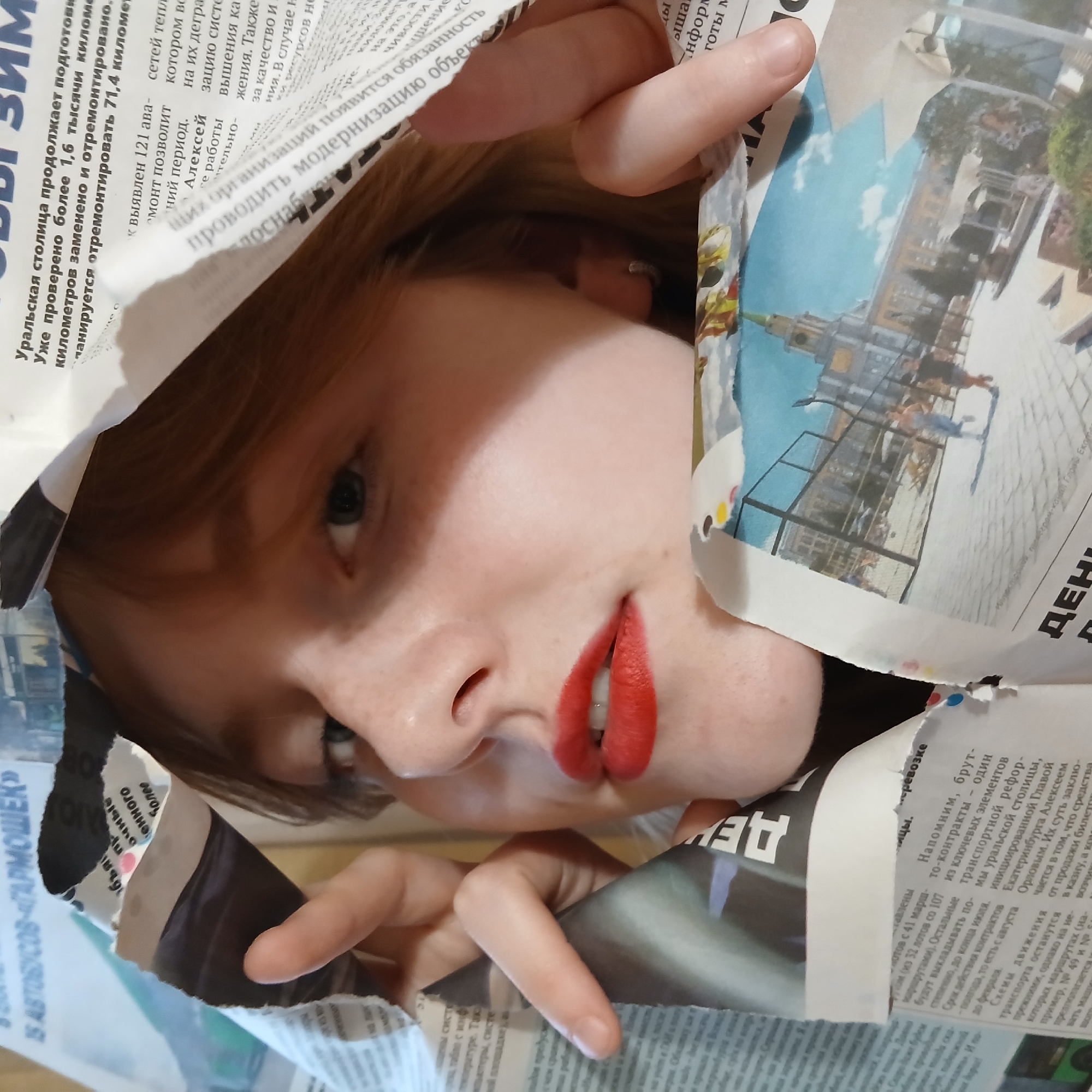 A young woman is depicted in a close-up portrait, framed by torn and crumpled sheets of Russian newspaper. Her face is angled slightly, with her right eye looking towards the viewer, and a subtle, enigmatic smile on her lips, accentuated by vibrant red lipstick. Her light brown hair is visible around her face, and her fair-skinned fingers emerge from and interact with the newspaper edges.
The newspaper, printed with black text and some color images depicting what appears to be a city scene with buildings and a blue sky, surrounds the woman. Visible text provides strong location cues, including "Уральская столица продолжает подготови" (Ural capital continues to prepare) and specific references to "Екатеринбурга Алексеем Орловым" (Yekaterinburg by Alexey Orlov), confirming the content relates to Yekaterinburg, Russia. Other discernible phrases mention "транспортной реформ" (transport reform) and "15 АВТОБУСОВ «ГАРМОШЕК»" (15 'accordion' buses), suggesting local news about urban infrastructure and development. The newspaper creates a unique, playful, and intimate framing for the subject. The time of day and weather are not discernible.