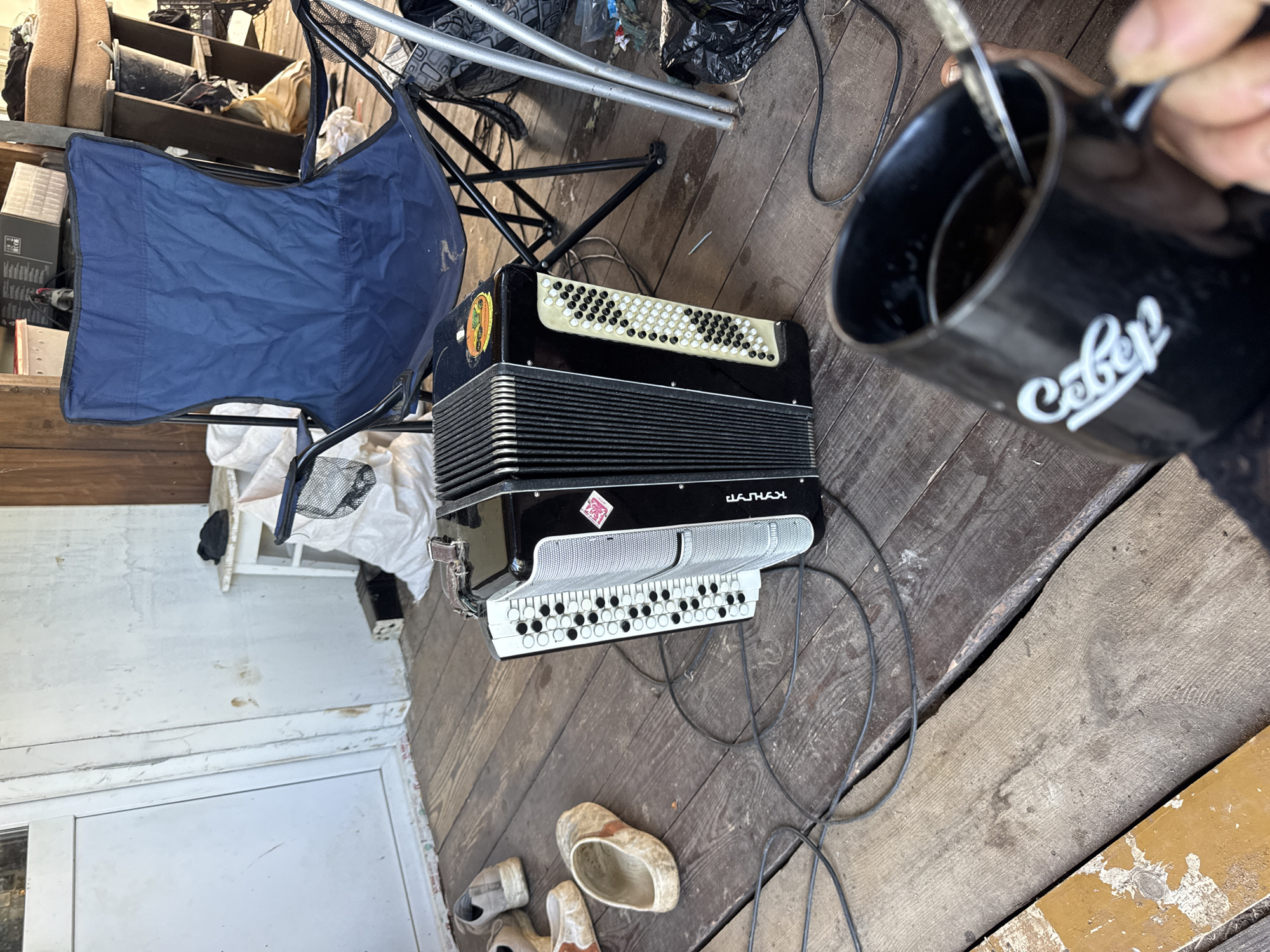 The image depicts an outdoor scene on a wooden porch, likely in the city of Chaikovsky, Russia, given the presence of an accordion with "КУНГУР" branding, a city in the Perm Krai region near Chaikovsky. In the foreground, a black mug filled with a dark liquid, possibly coffee, is being held by a person whose hand is visible. A spoon is inside the mug. To the left of the mug, an accordion rests on the wooden floor, connected by cables. Several pairs of shoes, including a pair of sandals and what appear to be sneakers, are scattered on the porch. Behind the accordion, a blue folding chair is set up. The background shows a white wall, a white wooden door, and various other objects, including bags, boxes, and shelving units, suggesting a cluttered, possibly storage area or an outdoor workspace. The overall impression is of a casual, perhaps break-time setting.