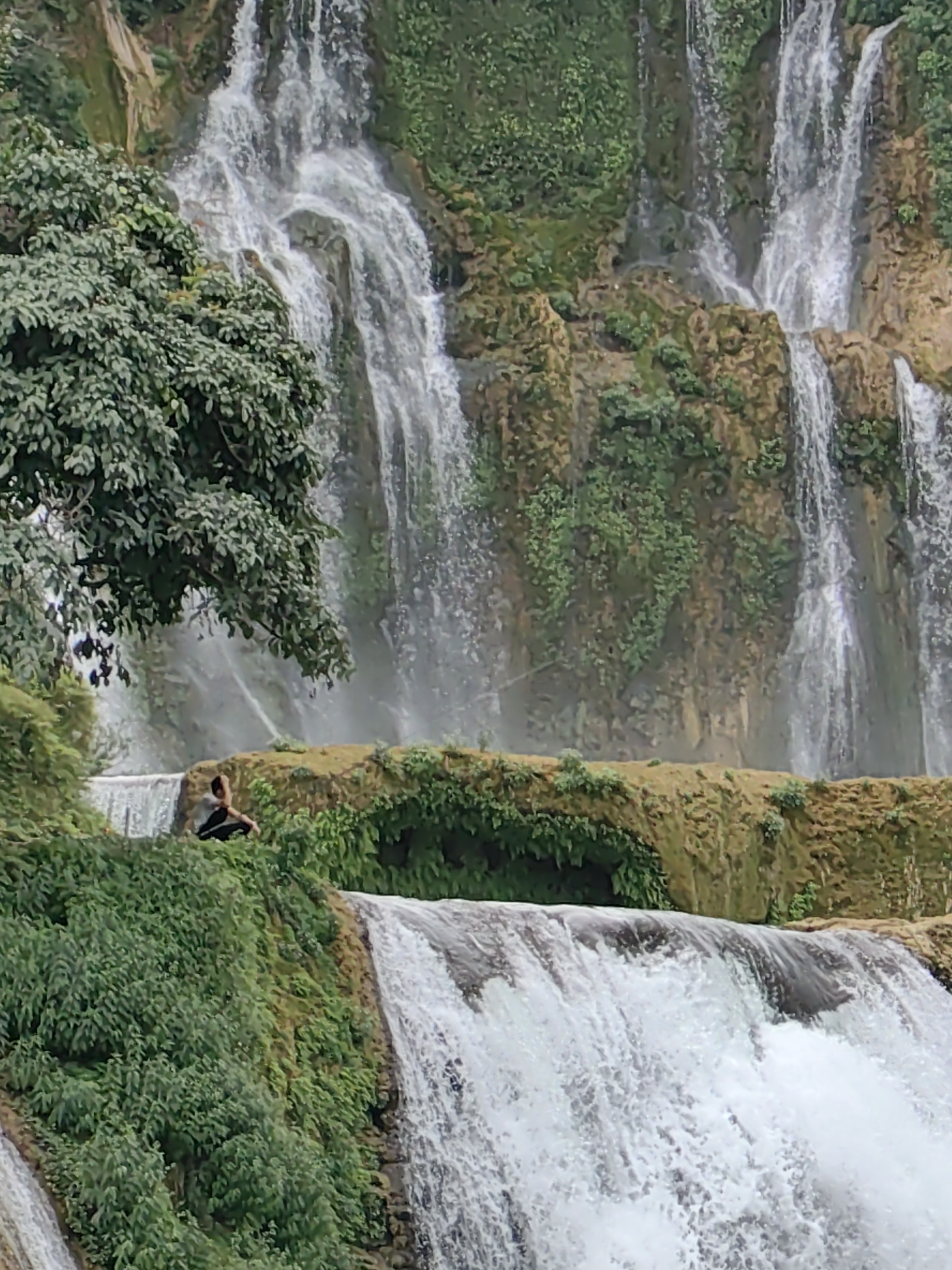 The image captures a scenic waterfall in Daxin County, China. The multi-tiered waterfall cascades down a mossy, rocky cliff covered in lush green vegetation. A lone individual is visible sitting at one of the levels along the waterfall.