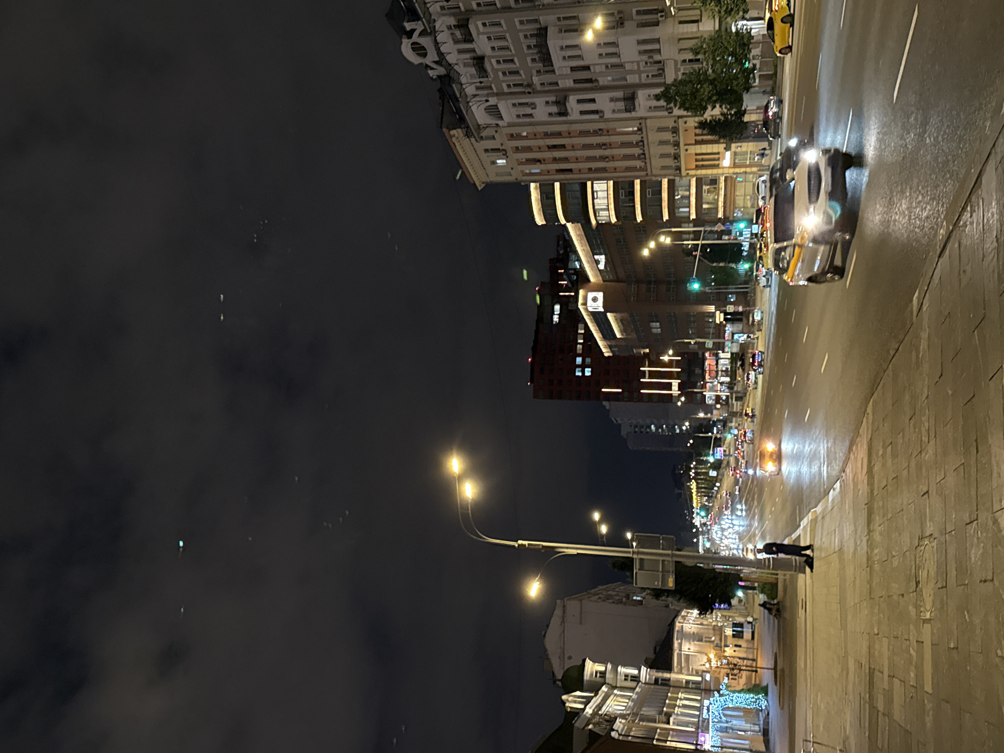 The image depicts a nighttime street scene in Moscow, Russia, likely on a wide avenue. In the foreground, a paved sidewalk is illuminated by streetlights, with a lone figure standing near a lamppost and a dark object on the ground. Across the street, traffic moves in both directions, with vehicle headlights and taillights creating streaks of light. Several multi-story buildings line the avenue, displaying illuminated windows and architectural details. The sky is dark and cloudy, with a few faint lights visible, possibly from aircraft or distant city lights. A white car with yellow and orange stripes, possibly a taxi, is prominent in the right lane, its headlights illuminated. The overall impression is that of a busy urban environment at night.