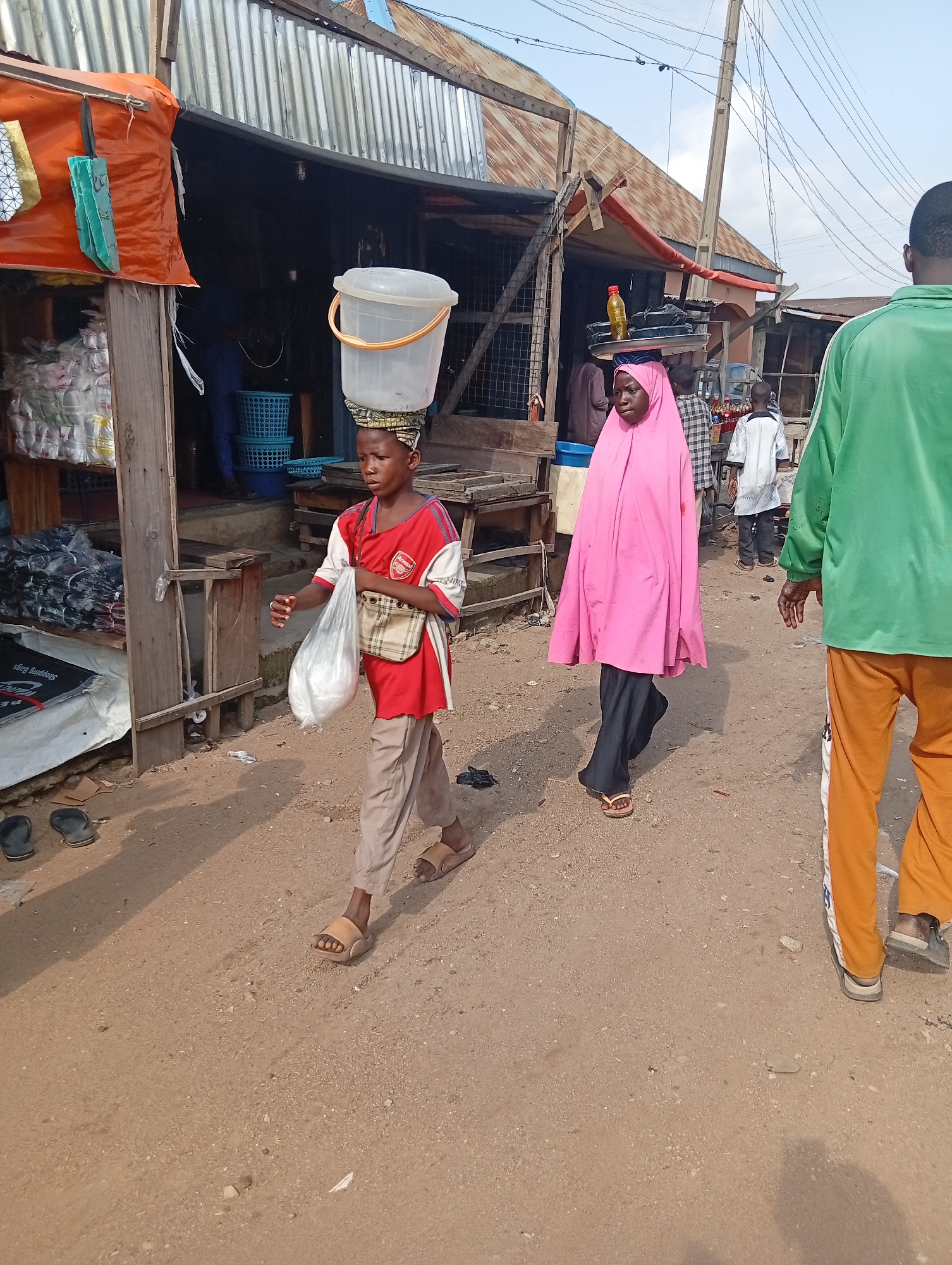 In Jalingo, Nigeria, a dusty market scene unfolds under a bright sky. A young boy, wearing a red and white Arsenal jersey and tan trousers, walks carrying a white plastic bucket on his head. He also holds a white plastic bag in his right hand. To his right, a young woman is seen wearing a pink hijab and dark trousers, with a tray of items balanced on her head. Behind them, a man in a green long-sleeved shirt and orange trousers walks away from the camera. The environment suggests a bustling marketplace with stalls and various goods displayed.