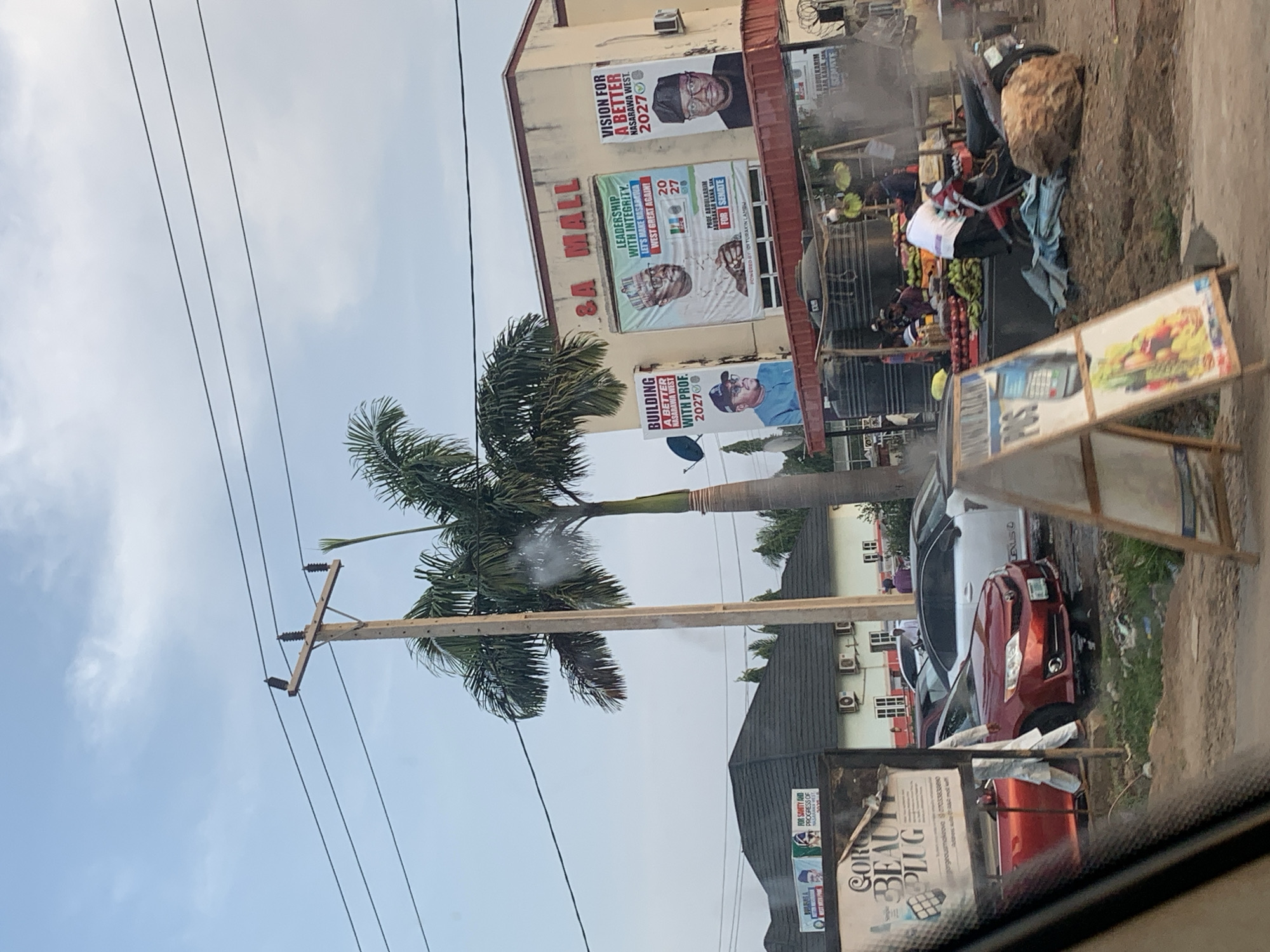 The image captures a street scene in Keffi, Nigeria. Buildings with campaign posters are visible, advertising candidates for the 2027 elections, with slogans like "Vision for a Better Nasarawa West" and "Building a Better Nasarawa West with Prof." The posters feature images of the candidates. A palm tree and utility pole are also present in the scene. On the street, a fruit stand with a variety of produce is visible, along with parked cars. A sign advertising "Coro Beauty Plug" is near a building. People are present near the fruit stand. The photo is taken from inside a vehicle, looking out onto the street.