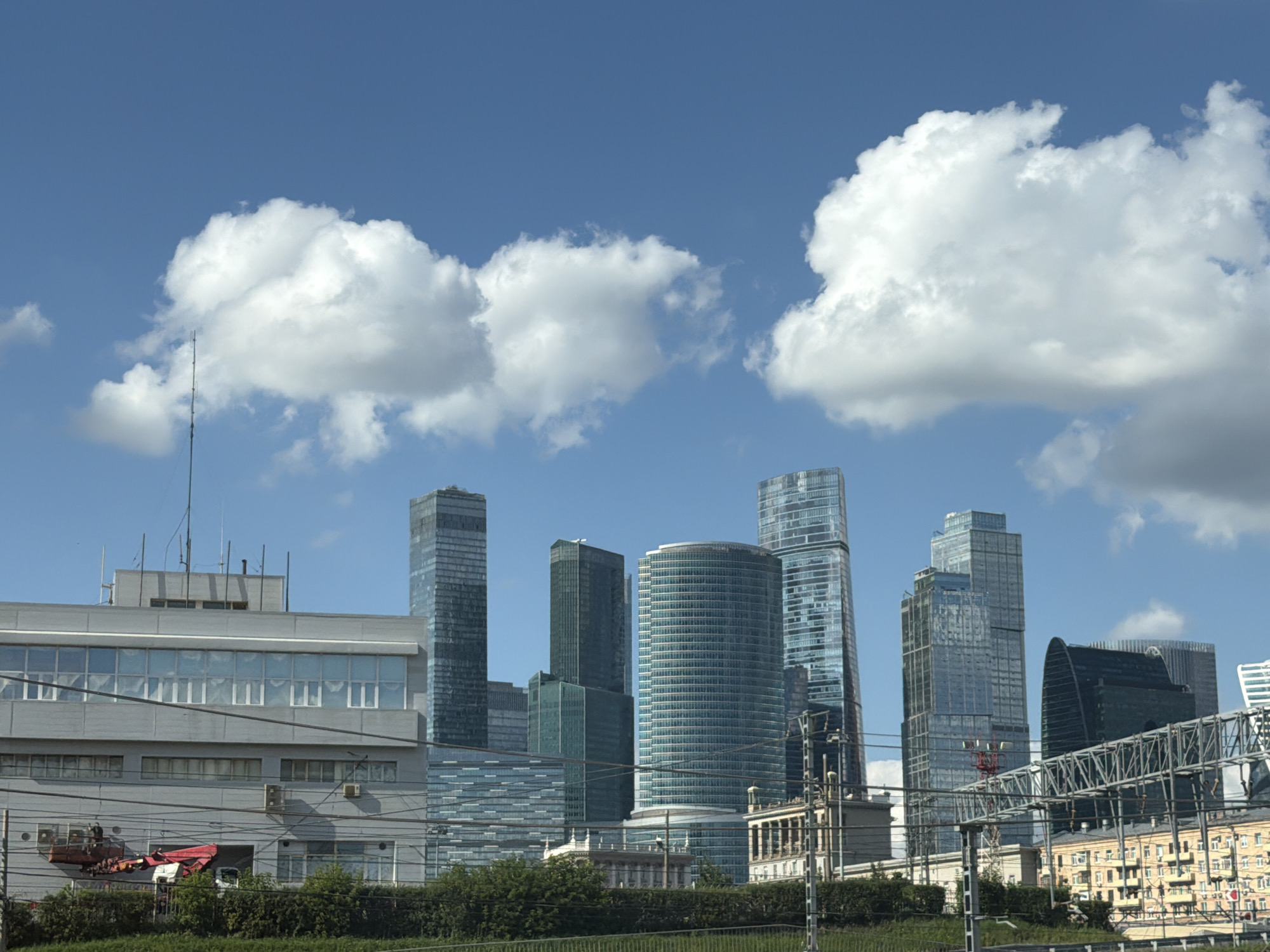 This image captures a view of the Moscow International Business Center, also known as Moscow City, in Moscow, Russia. The foreground features a low-lying, gray building with multiple windows and visible electrical wires running in front of it. A red and white crane is partially visible on the left side of this building. Behind this structure, a cluster of modern skyscrapers dominates the skyline, characterized by their glass facades and varying heights and architectural styles. Prominent among them is a tall, slender skyscraper with a blue-tinted glass exterior, and a distinctive curved, dark-green building. The sky above is a bright blue, dotted with large, fluffy white clouds. The overall scene depicts a contemporary urban landscape with a mix of functional and iconic architecture against a clear sky.