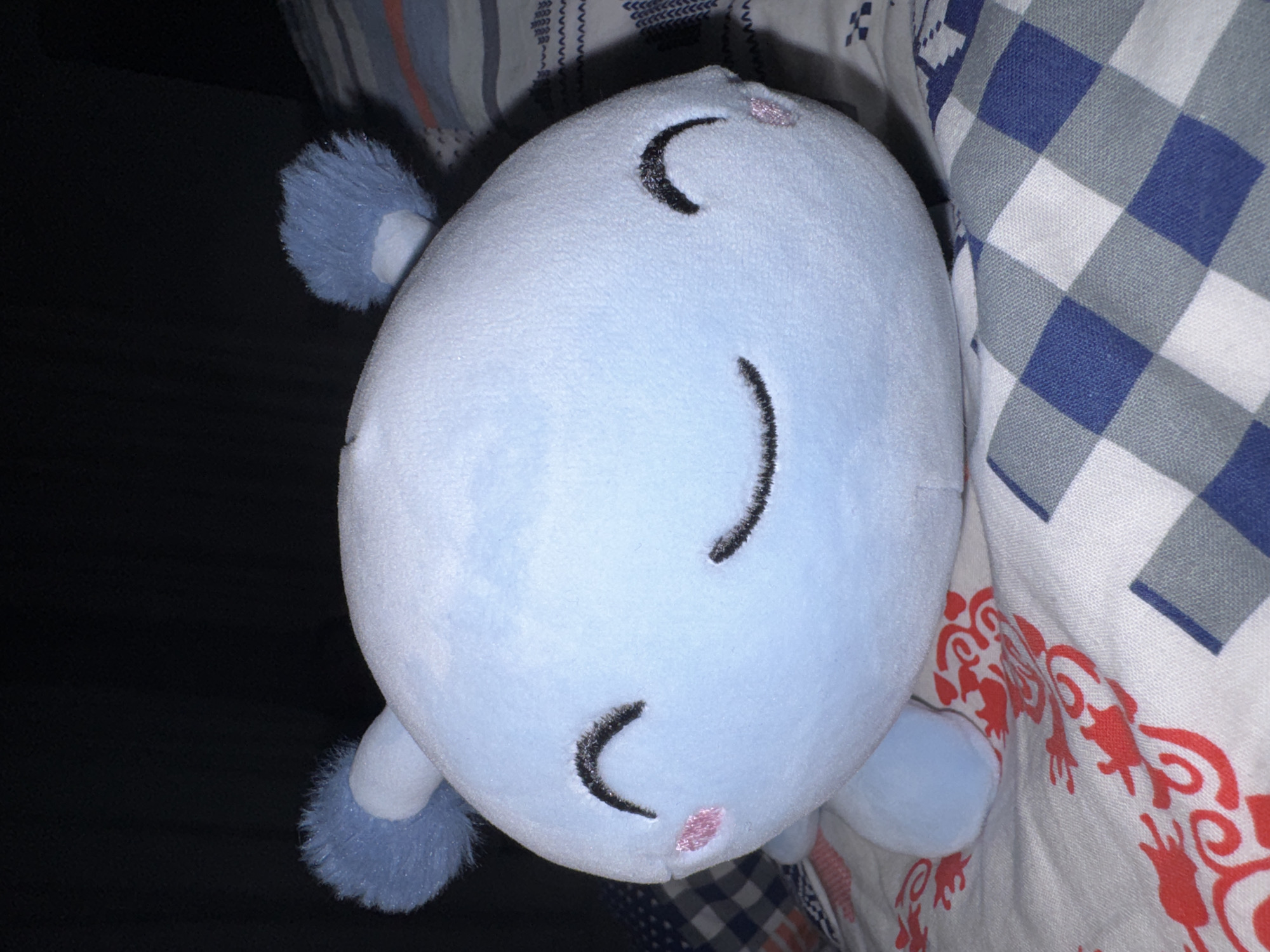 The image displays a light blue, oval-shaped plush stuffed animal, oriented horizontally. The stuffed animal features embroidered black crescent-shaped lines representing closed eyes and two small pink embroidered dots below them, suggesting a sleeping or peaceful expression. Small, fuzzy blue appendages, possibly limbs or ears, are visible extending from its sides.
The stuffed animal rests on or is surrounded by various patterned textiles. To its right, there is a white fabric with a blue and gray checkered pattern. Below the stuffed animal, a white fabric with an ornate, flowing orange-red design is visible. In the upper left background, striped fabric in blue, white, and possibly red, along with a darker fabric featuring a white geometric pattern, can be seen. The area to the left of the stuffed animal is dark, resembling a deep shadow or a plain dark surface.
The lighting appears dim, suggesting an indoor setting, possibly with low ambient light. No specific activity is depicted other than the stuffed animal resting. There is no visible text in the image.