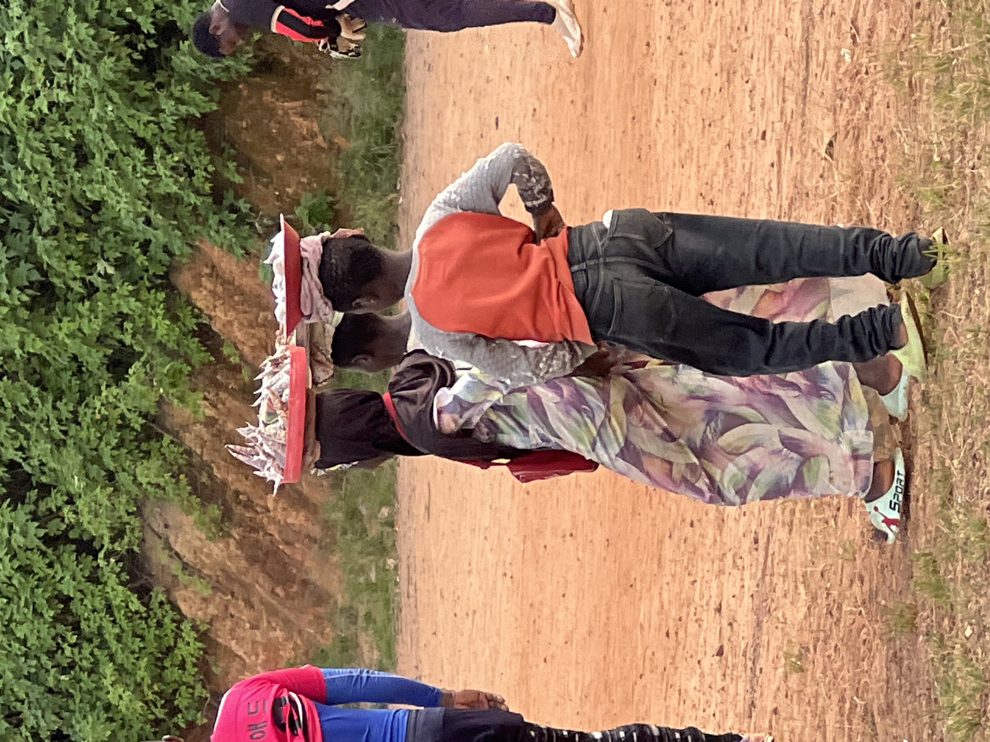 In Jos, Nigeria, on a dirt path beside a grassy verge and a steep, eroded hillside covered in dense green foliage, two young individuals, likely of West African descent, are walking. The person in front is wearing a long, flowing dress patterned with purple and green watercolor-like splotches, and a dark hijab. On their head, balanced precariously, is a red plastic tray piled high with small, white items, possibly dried fish or snacks. Another tray is visible on the head of a third person, partially obscured, also carrying similar items. To the right, a boy in a grey long-sleeved shirt with an orange yoke and dark jeans stands with his hands on his hips, facing away from the viewer. The foreground is a reddish-brown dirt ground scattered with sparse green grass. In the background, a person in a black and red shirt is visible in the distance.