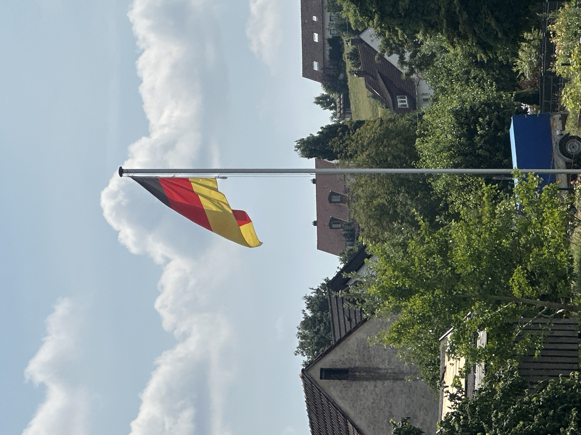 A German flag flies on a flagpole against a backdrop of a blue sky with scattered white clouds. The flag, positioned on the left side of the frame, displays the national colors of black, red, and yellow. In the lower half of the image, several houses with dark tiled roofs are visible, nestled among lush green trees. The architecture suggests a residential area, possibly in a suburban or rural setting within Günzburg, Germany. One house on the left is a light-colored stone structure with a dark, rectangular opening. Another house in the middle ground has a dormer window. In the foreground, on the bottom left, part of a dark fence is visible, and a tree with bright green leaves occupies the center. To the right of the flag, a small blue trailer is parked. The scene appears to be a sunny day.