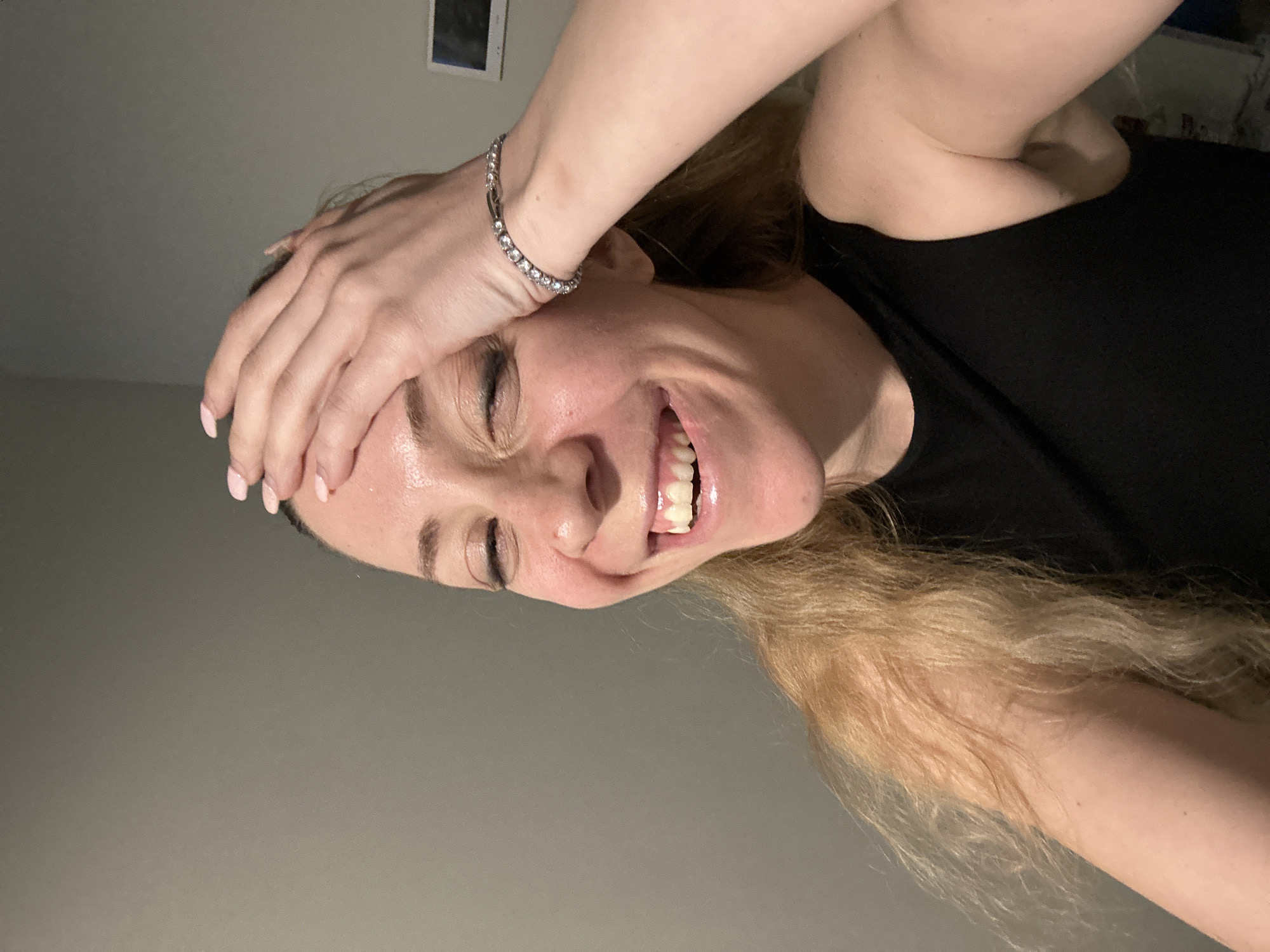 A woman is pictured close-up with her face tilted towards the camera. She is fair-skinned, smiling widely, and has blonde hair. Her right hand is resting on her forehead, adorned with a silver bracelet. She is wearing a black shirt. The background is a plain light grey wall with a small dark framed object hung on the wall above. There is no indication the image was taken in Amsterdam, Netherlands.