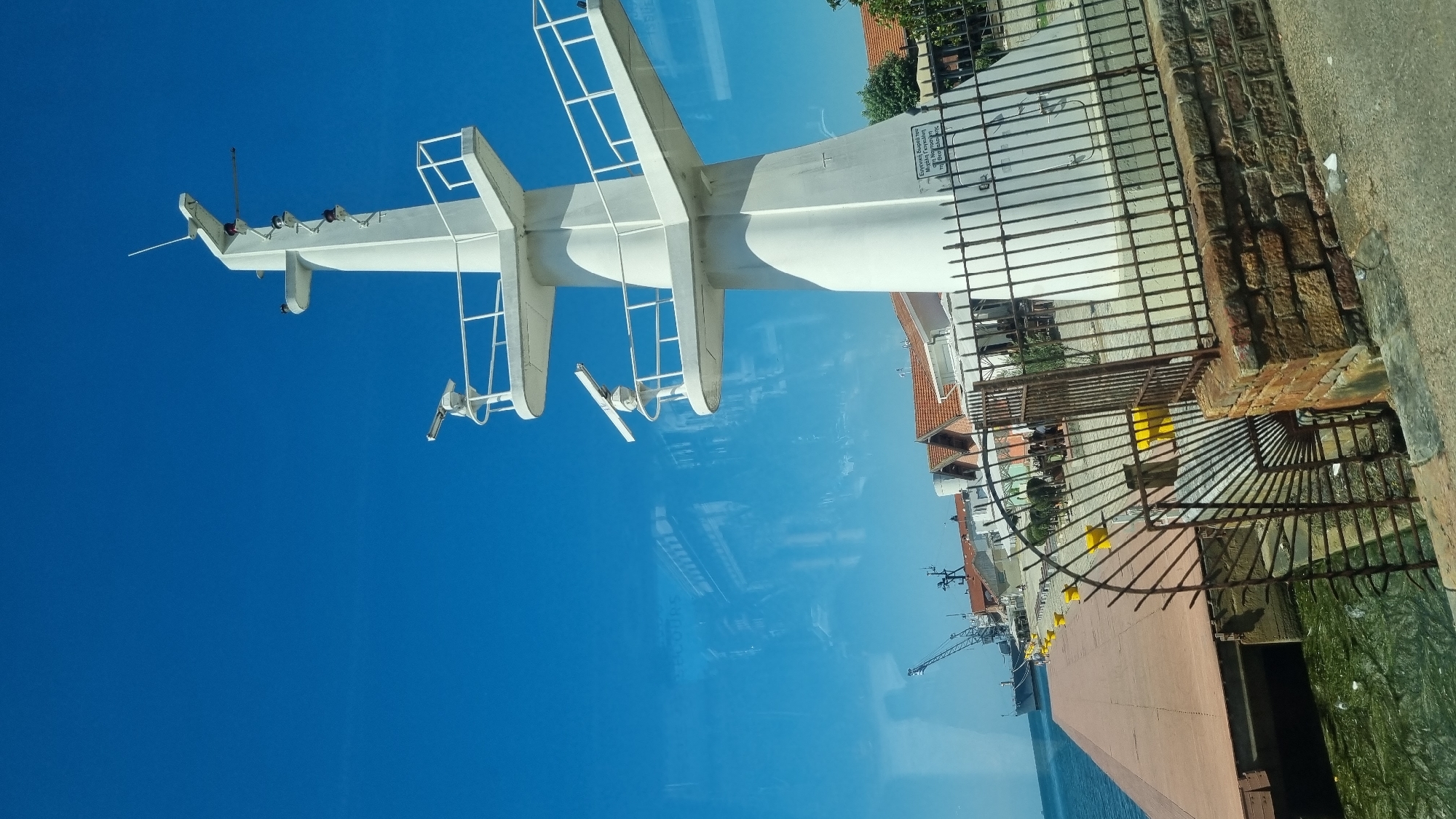 This image captures a view of the waterfront in Thessaloniki, Greece, focusing on a prominent white mast structure with radar equipment. The mast stands tall against a clear blue sky and is situated near a dock. In the foreground, a weathered metal fence and brick wall partially obscure the view of the dock. The dock itself is constructed of reddish-brown planks and leads to the water, which appears somewhat murky with green algae.
Further along the dock, several yellow buoys are visible, and buildings with orange-tiled roofs line the background. A large ship and a crane are docked in the distance, suggesting a functioning port. Reflections on a glass surface in the foreground distort the image, showing glimpses of buildings and the sky. Some indistinct figures are present on the dock area. The mast itself has multiple platforms with railings, and at least two radar dishes are mounted on it, along with several red lights. A sign with Greek text is affixed to the base of the mast. The overall scene depicts a typical port environment under a bright, sunny sky.