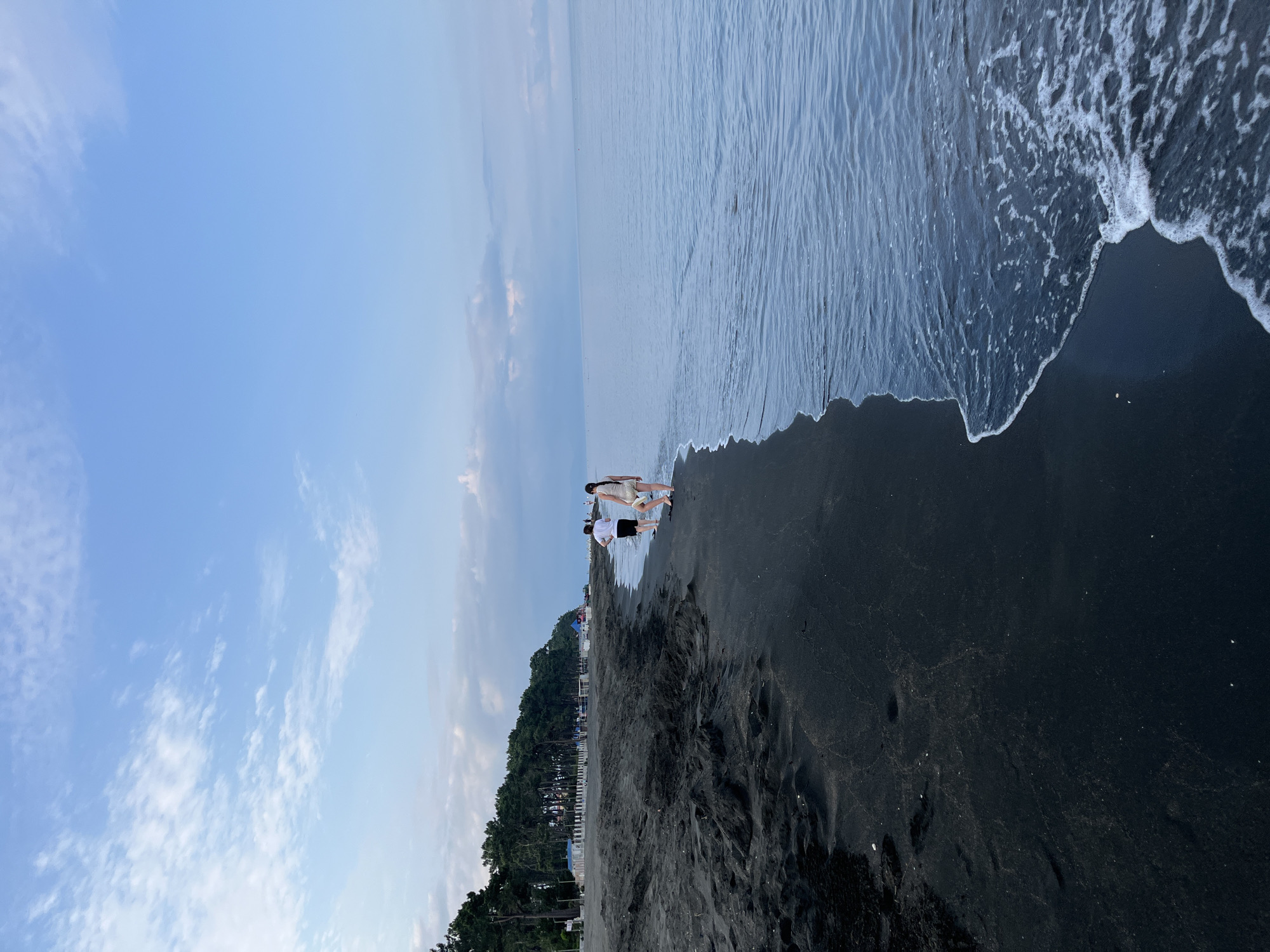 In the city of Ureki, Georgia, two people are walking along a black sand beach. The man is wearing a white t-shirt and black shorts, while the woman is in a light-colored dress. The sea is calm with gentle waves lapping at the shore. In the background, a line of pine trees borders the beach, and a few buildings are visible, suggesting a developed area along the coastline. The sky is blue with scattered white clouds, indicating fair weather. The sand is dark and appears somewhat wet near the water's edge, with some textured patterns formed by the receding tide.