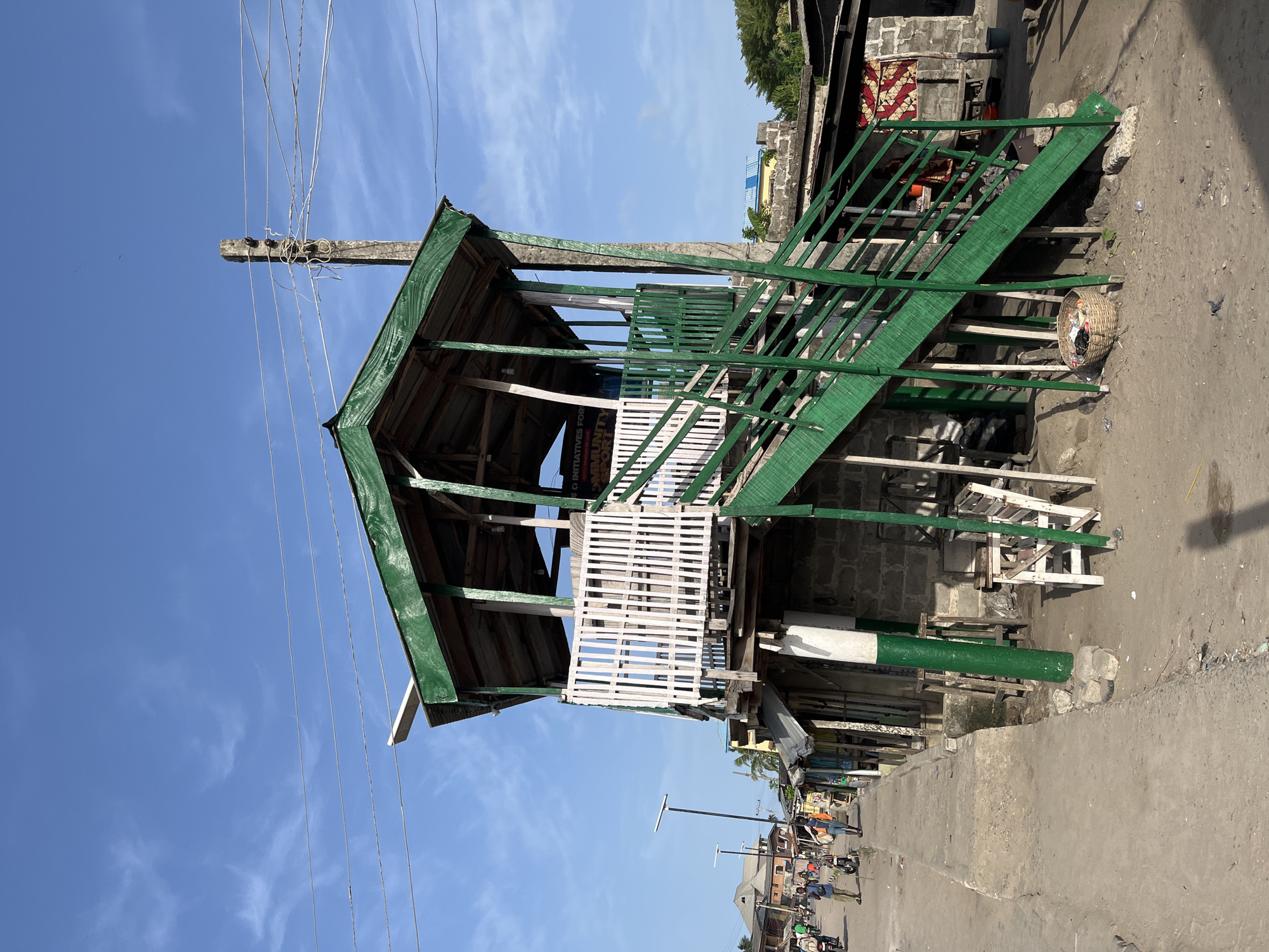 The image shows a two-story elevated wooden structure in Badagry, Nigeria. The structure has a green roof, green-painted metal stairs, and white latticework. Power lines are visible above the structure, connected to a wooden pole. Below the structure, there's a concrete pavement or sidewalk. People and motorcycles are seen further down the street. The sky is blue with scattered clouds.