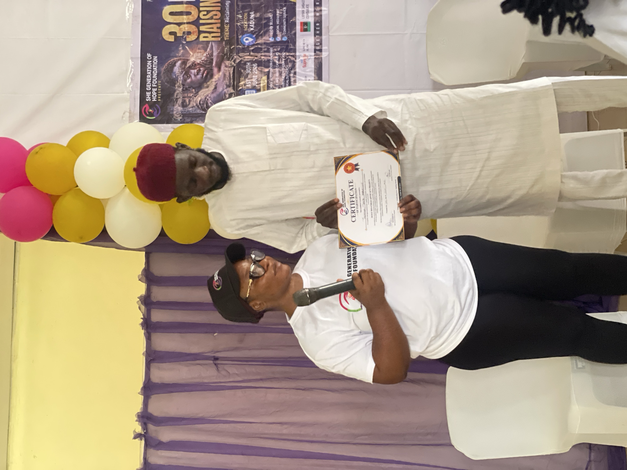 In Unguwar Shanu, Nigeria, a woman in a white t-shirt and black leggings, wearing glasses and a black cap, stands on the left holding a microphone and a certificate. To her right, a man in a white traditional outfit and a red cap is also holding the certificate, which is being presented. Balloons in pink, yellow, and white are visible in the background, along with purple curtains and a banner that reads "SHE GENERATION OF HOPE FOUNDATION PRESENTS 30 R." The scene appears to be an indoor event, possibly a presentation or award ceremony, taking place in a room with white chairs.