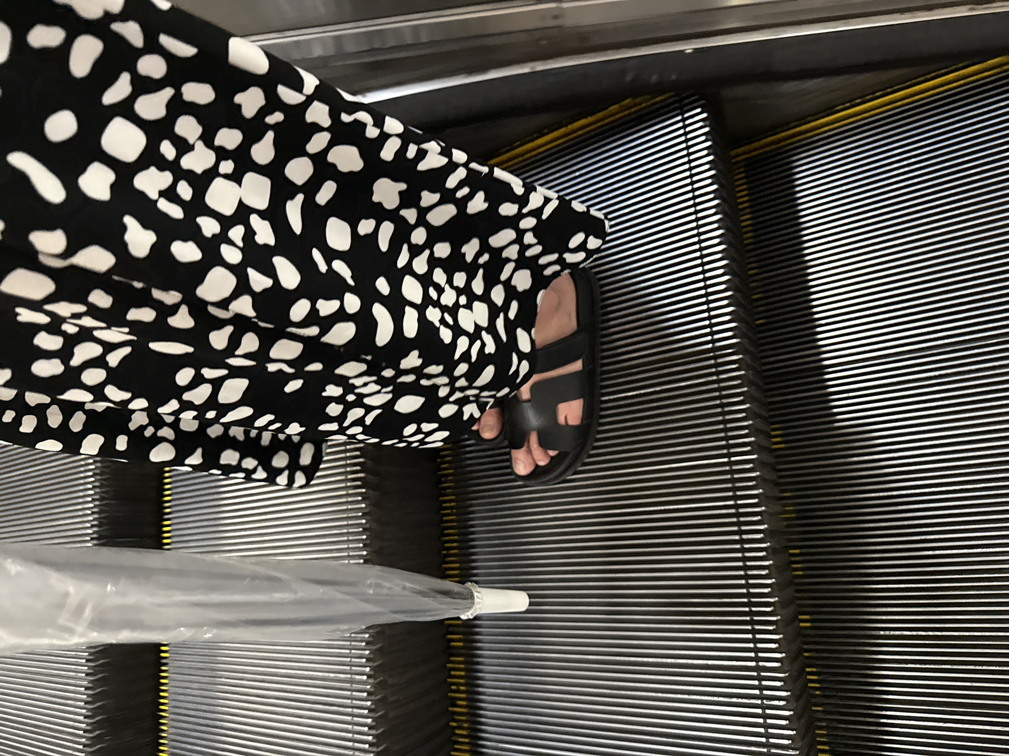 A person stands on an escalator in Haizhu District, China. The person is wearing a black and white spotted dress and black sandals. They are holding a clear plastic umbrella in their left hand. The escalator steps are metallic with yellow safety strips. The image is taken from a low angle, focusing on the lower half of the person and the escalator.
