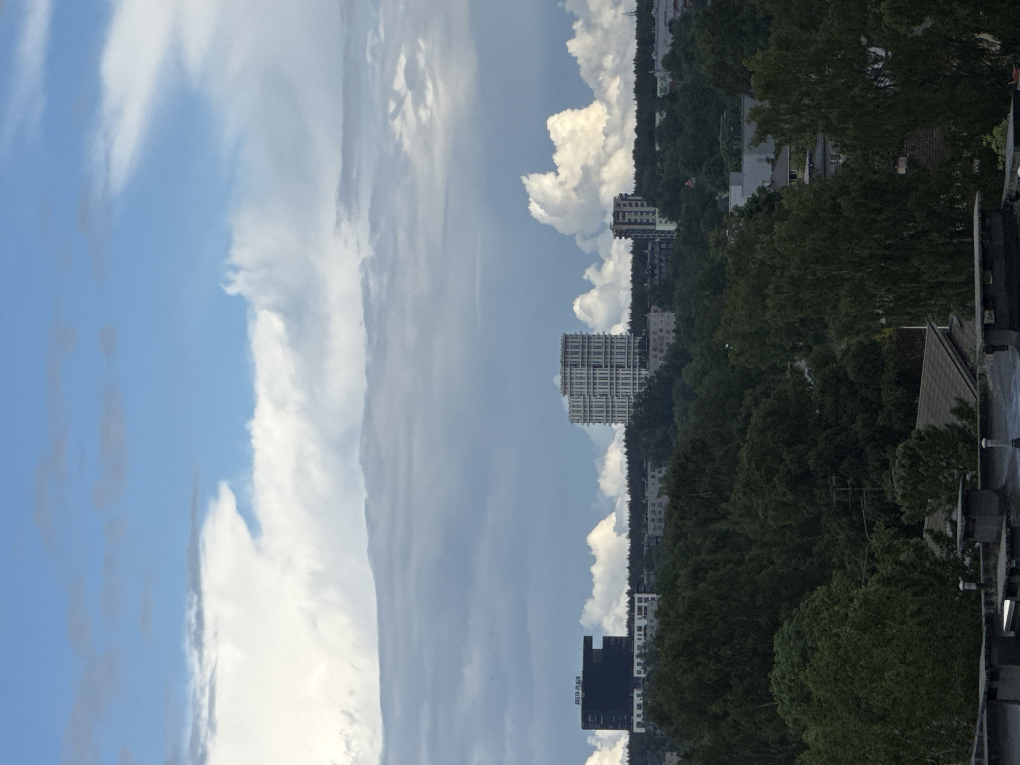 The image captures an urban landscape in Tallinn, Estonia, under a partially cloudy sky. In the mid-ground, a dark, rectangular building with the text "DELTA PLAZA" is visible on its left side. To its right, a white, multi-story building with a distinctive, stacked architectural design stands out. Further in the distance, another tall, narrow building is situated. The foreground features the rooftops of residential buildings, characterized by grey roofing and visible chimney structures. The mid-ground is dominated by dense green trees, forming a thick forest that separates the foreground from the city's buildings. The sky is a mix of bright blue patches and large, dramatic white and grey clouds, suggesting dynamic weather conditions. No people or immediate actions are discernible in the scene.