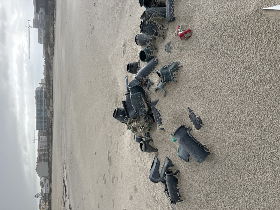 The image depicts a sandy beach in Gafanha da Nazaré, Portugal, under an overcast sky. In the foreground, a prominent cluster of approximately 20-30 dark grey, irregularly shaped plastic or ceramic fragments, consistent with broken containers or fishing gear components, lies partially buried in light tan sand. These pieces exhibit varying cylindrical and cup-like forms, many showing signs of wear and marine exposure. Entangled within the central part of this debris pile are strands of white string, dark green netting, and other fibrous material. To the right of the main debris, a crushed red and silver metallic object, resembling a discarded beverage can, is partially visible. The midground shows an expanse of fine-grained sand with subtle ripple patterns and scattered footprints. No individuals are present on the beach. In the background, a row of urban structures, including multi-story buildings with modern designs and glass facades, line the horizon. Between the beach and the buildings, sand dunes with sparse vegetation and a wooden walkway system are visible. Two large construction cranes are distinguishable against the grey, cloudy sky, indicating ongoing development behind the beachfront structures.