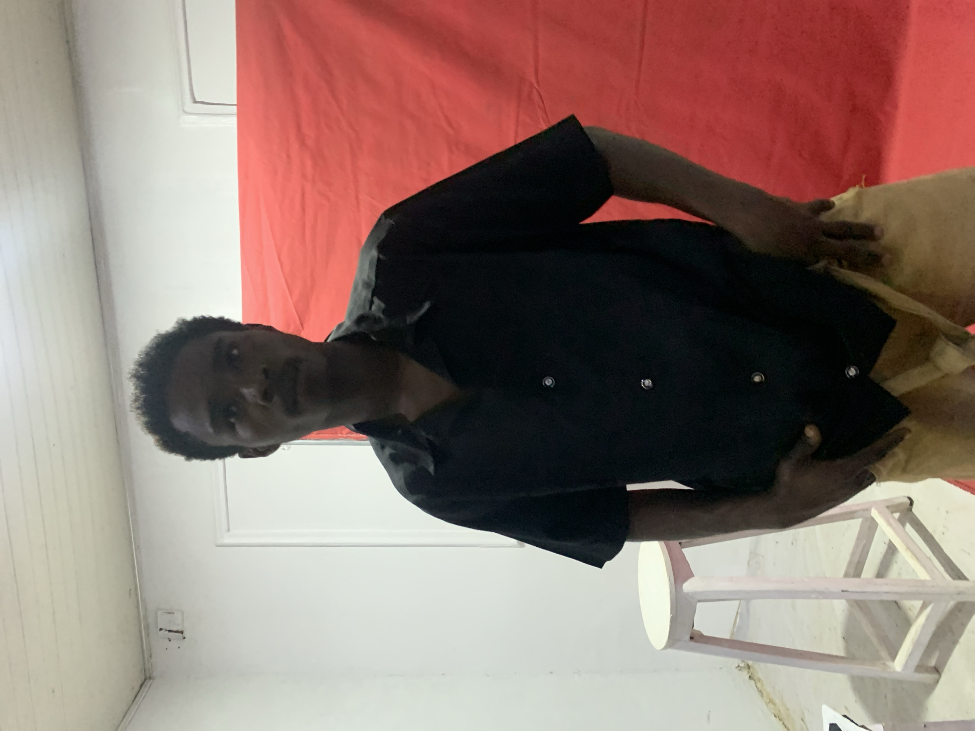 A man with dark skin and short, dark hair is indoors, wearing a black short-sleeved button-down shirt and tan pants. He stands near a wooden stool with a round white top and light pink painted legs. Behind him is a red fabric backdrop. The surrounding environment includes white walls and ceiling. The image does not contain enough information to determine if it is in Jos, Nigeria.