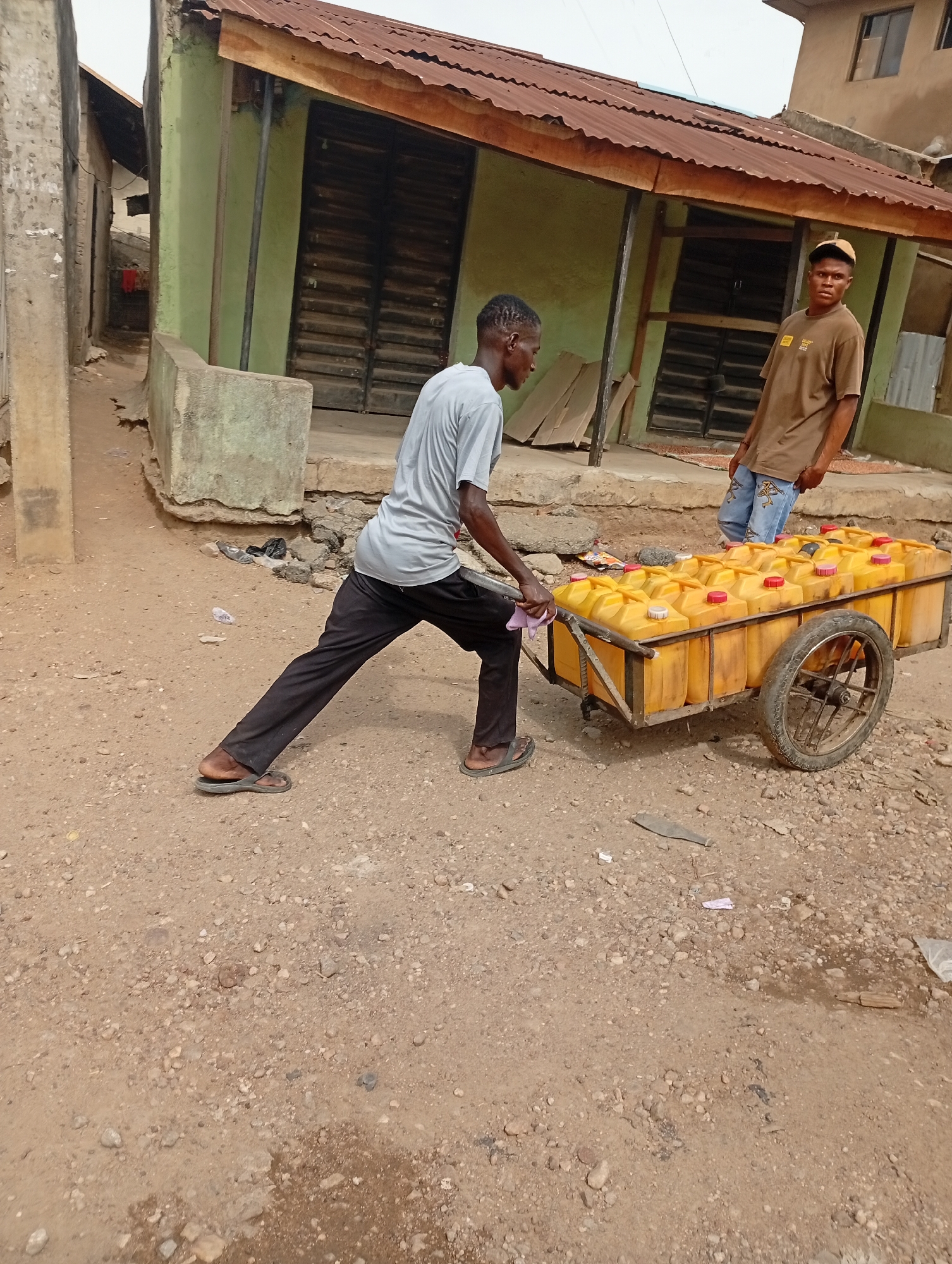 In Adankolo, Nigeria, a young man in a light gray t-shirt and black pants pulls a two-wheeled cart loaded with yellow jerrycans, likely filled with water or fuel. He wears sandals and leans forward, exerting effort to move the heavy load. Another young man, wearing a brown t-shirt, blue jeans, and a tan baseball cap, stands near a building and observes. The setting is a dirt road lined with buildings of varied construction. The foreground shows a dirt and pebble surface.