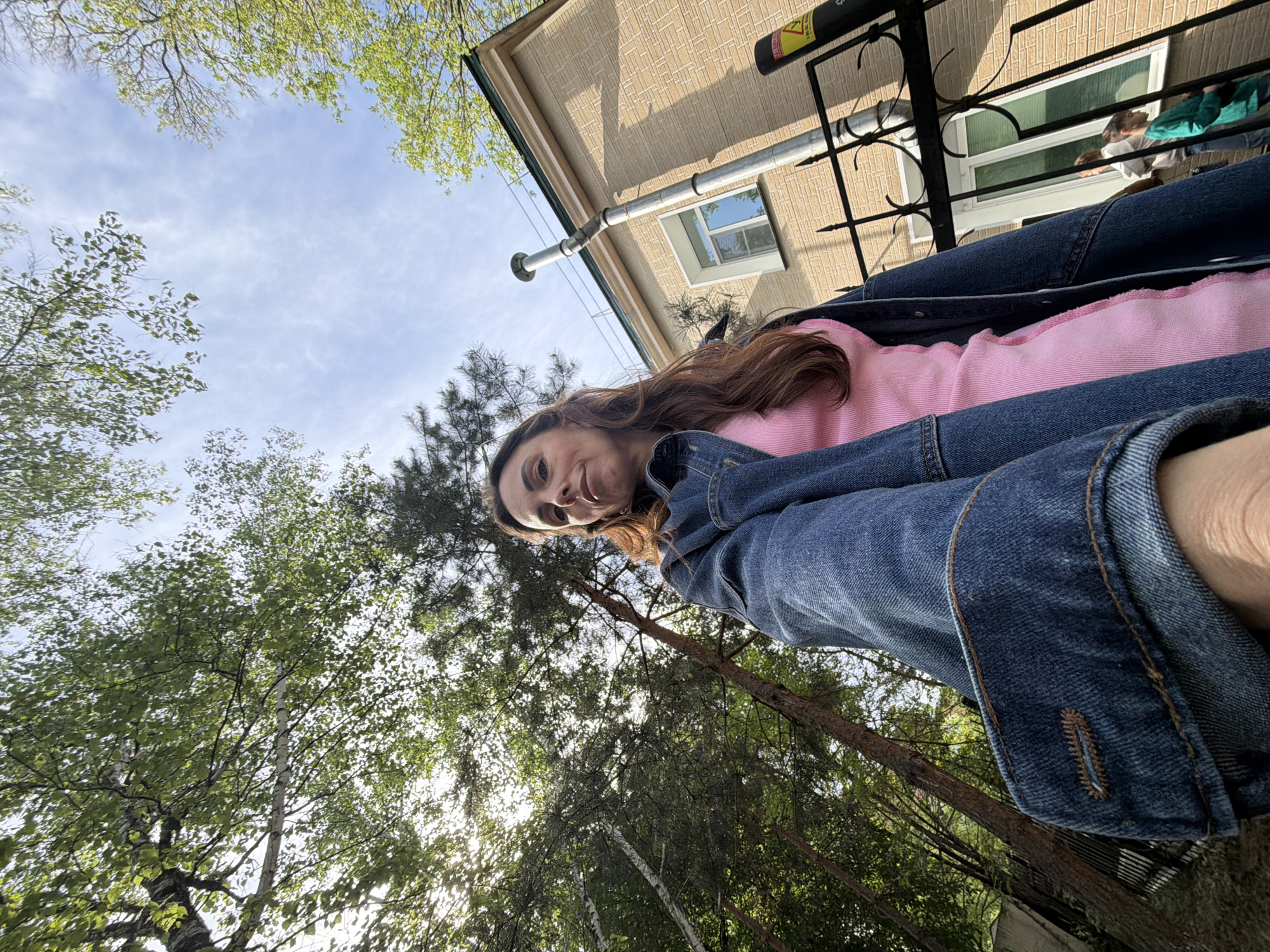 A low-angle shot captures a woman in a pink top and denim jacket, seemingly taking a selfie. Above and behind her, a building with beige siding and a black wrought-iron balcony is visible. On the balcony, other people are present. To the left and below the woman, trees with green leaves fill the frame, with a glimpse of a cloudy sky. The scene appears to be outside, possibly a backyard or park. There is no indication in the image that the location is Birobidzhan, Russia. The location is unknown.