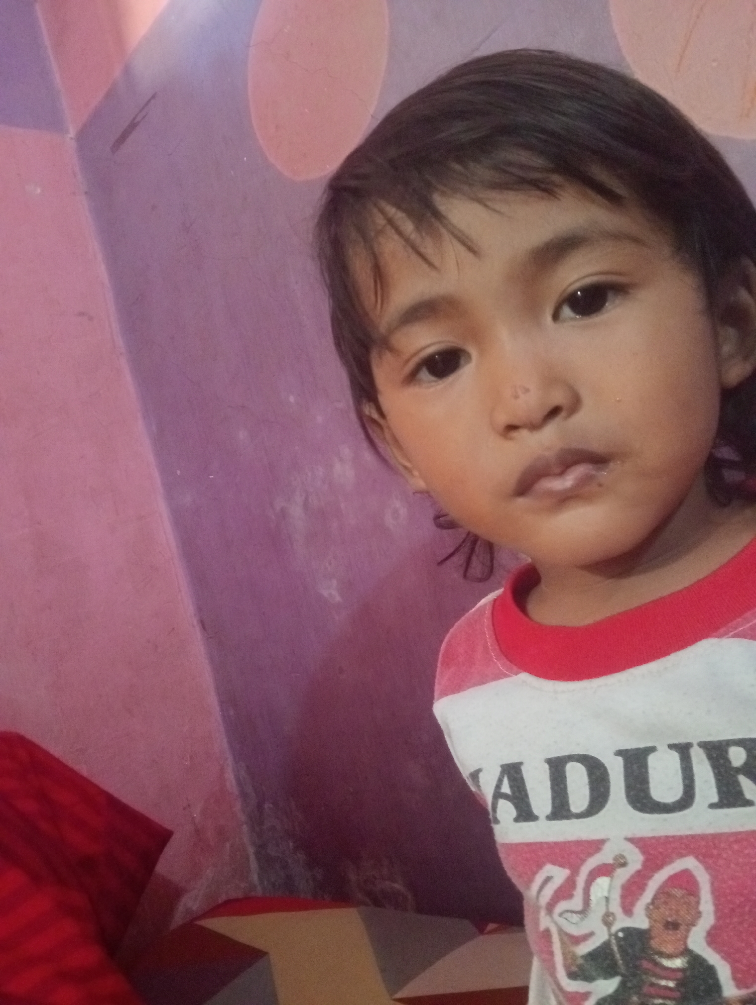 This media file is a close-up, waist-up portrait of a young Indonesian child, likely a girl, against a backdrop of a wall. The child has dark hair and appears to be looking directly at the camera with a neutral expression. Her shirt is visible, featuring a red collar and a white band with dark lettering. The visible text reads "MADUR" followed by an image of a person in traditional attire. The wall behind the child is painted in two shades of purple and pink, with some circular patterns visible. The lighting suggests it is daytime. The scene is set in Bloro, Indonesia.