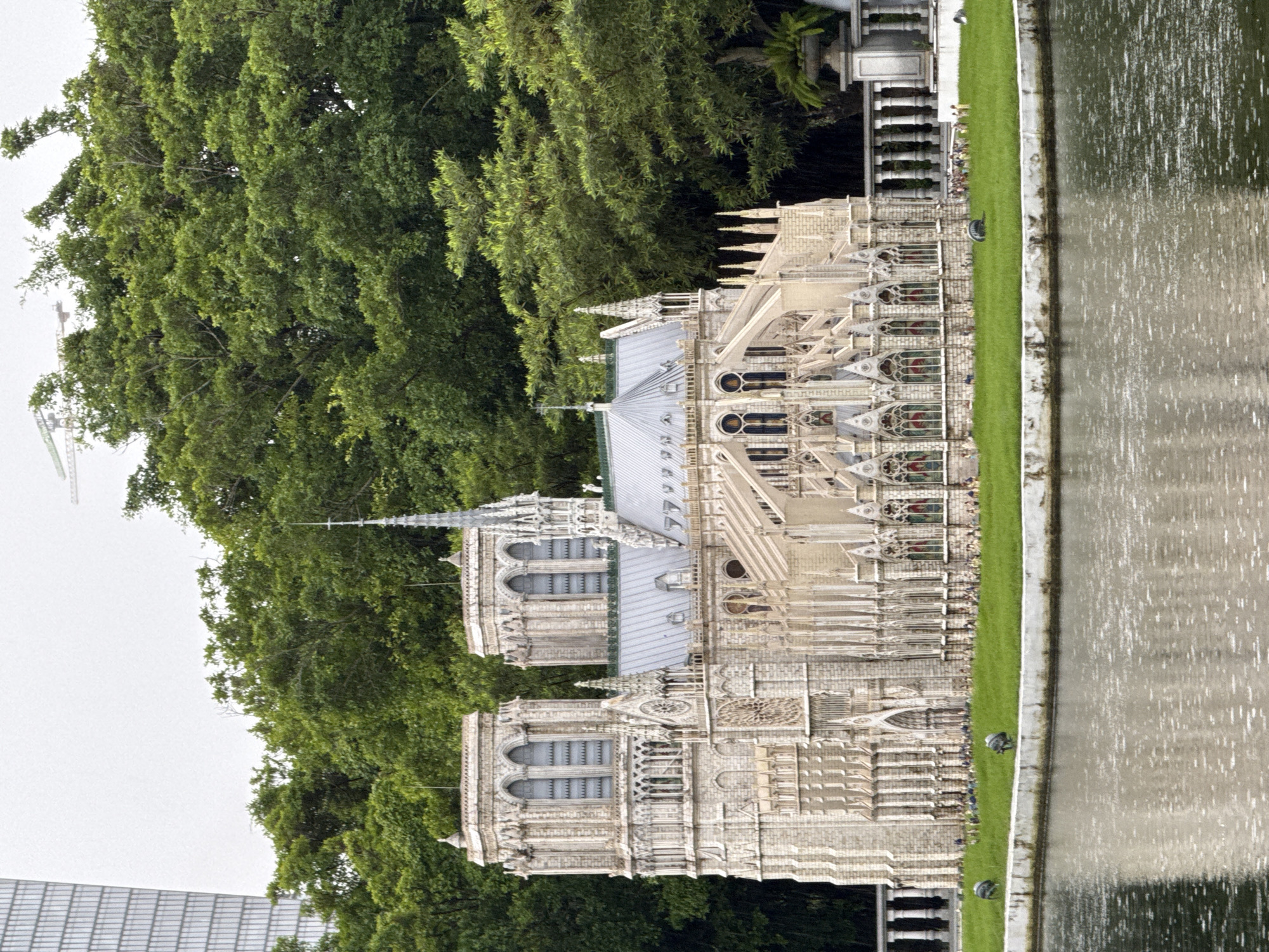 The image shows a miniature replica of Notre Dame Cathedral situated on a small island surrounded by water, likely part of the Splendid China Folk Village in Nanshan District, China. The replica is detailed, featuring Gothic architecture with spires, arched windows, and intricate stonework. Lush green trees form a backdrop to the miniature cathedral, with a modern high-rise building visible in the far left background. The water in the foreground reflects the sky and the surrounding greenery. The lighting suggests an overcast or diffused daylight condition. There are no visible people in the immediate vicinity of the miniature, but small figures appear to be placed along the grassy area in front of the structure, suggesting it's a display for visitors. The overall scene depicts a tourist attraction with a focus on architectural miniatures and cultural representation.