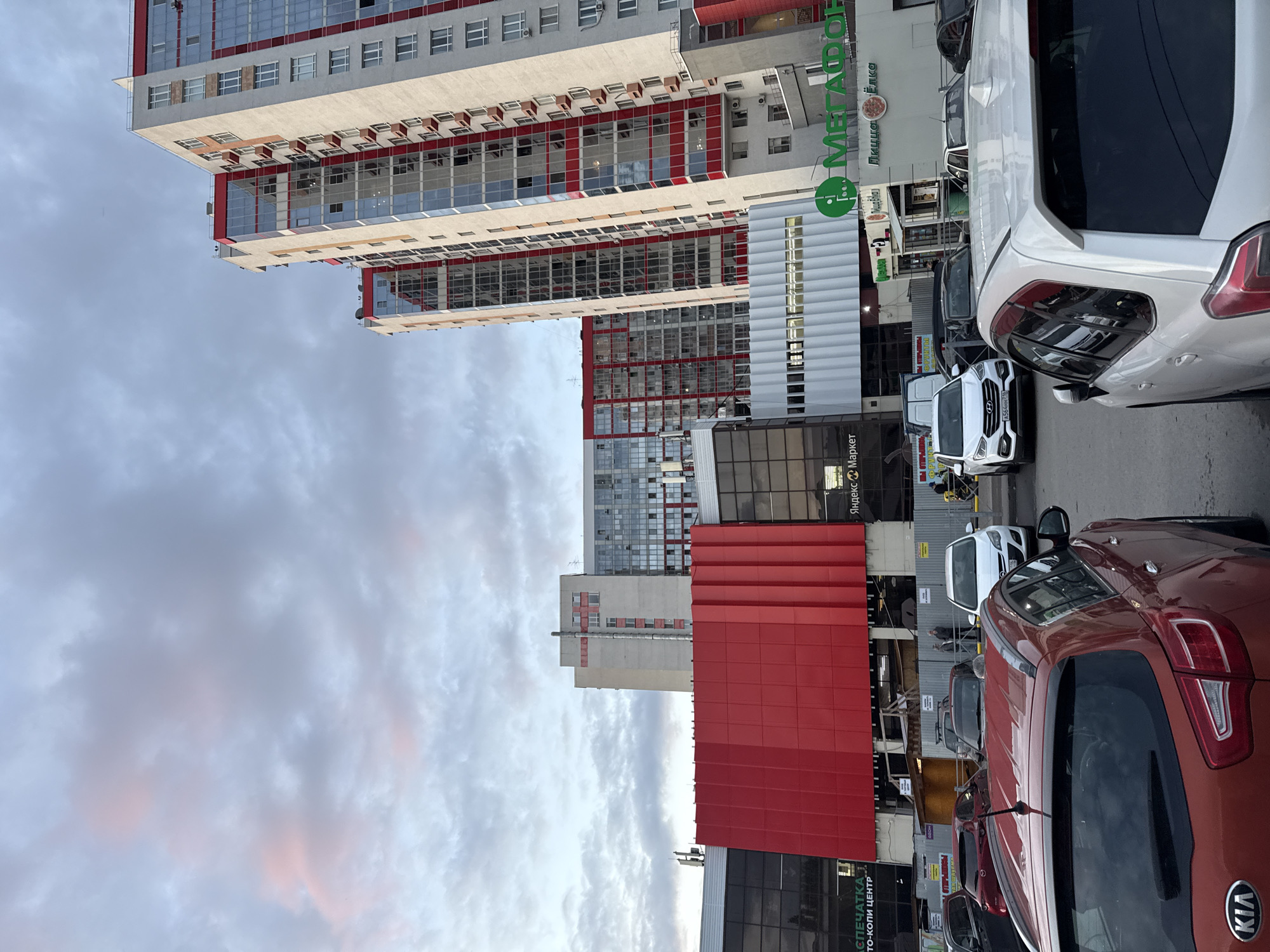 The image captures a street-level view of a modern urban scene in Kazan, Russia. In the foreground, several cars are parked on a paved surface, including an orange Kia SUV and a white Subaru SUV. Behind the cars, a multi-story building complex dominates the background, featuring a large red facade on one section and tall, contemporary residential buildings with prominent red and white accents and numerous glass windows. The sky above is overcast with diffused light, hinting at an early evening or late afternoon setting. Signs indicating businesses such as "Яндекс Маркет" and "Мегафон" are visible on the lower levels of the building complex, suggesting a commercial and residential mixed-use area. The overall impression is of a bustling city environment.