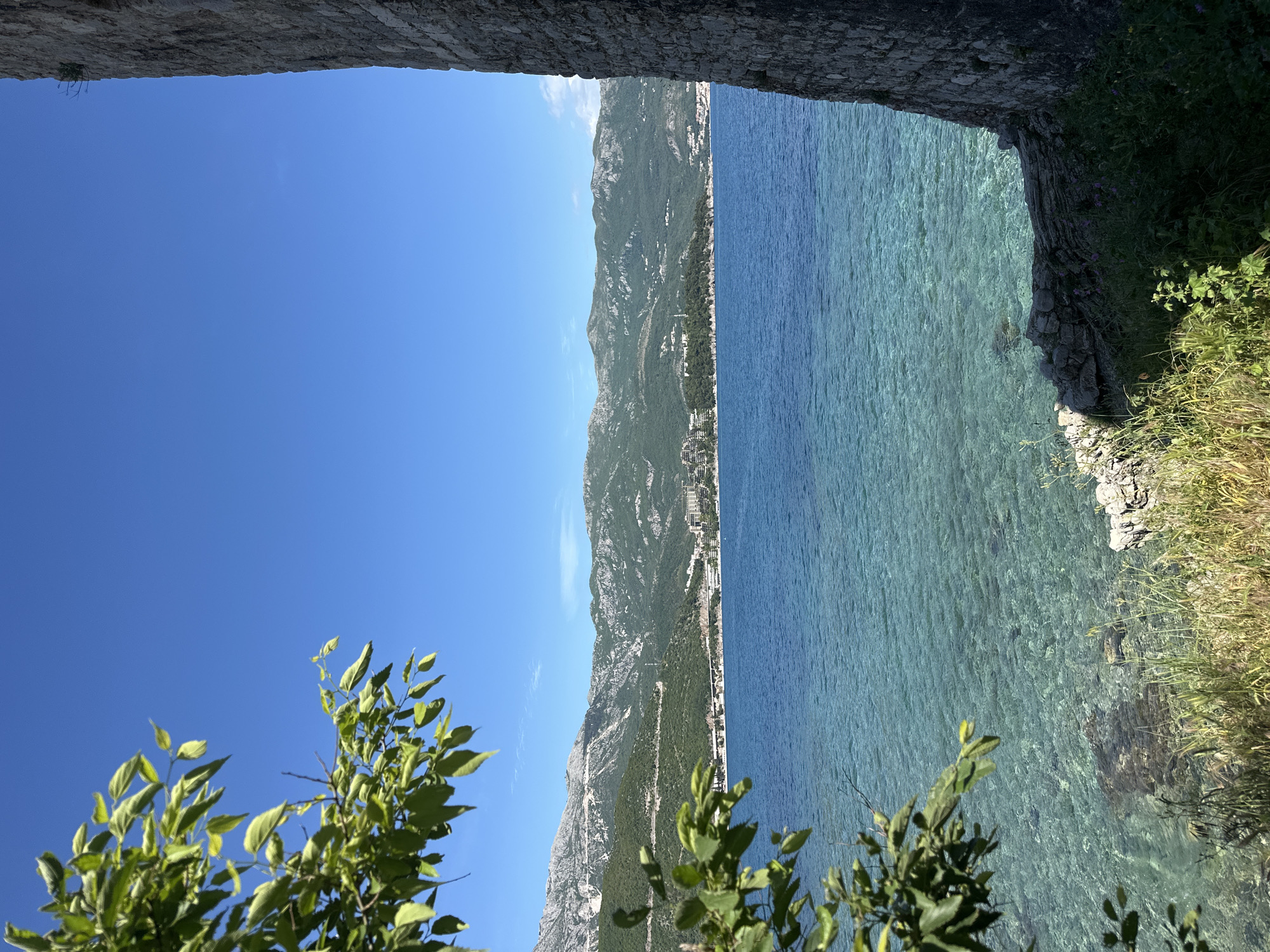 The image is an outdoor shot from what appears to be a fortified structure, perhaps a castle wall, framing a view of the Adriatic Sea and coastal mountains. The view is taken from a high vantage point, looking toward the sea. The sky is clear blue and takes up a large portion of the left side of the frame, offset by some foliage. The mountains stretch along the coastline in the background, a city strip is nestled between the mountains and the sea. The sea itself is calm and clear, with visible underwater rocks and details near the shore. The foreground includes parts of the stone structure from which the photo is taken, with some vegetation growing on the rocks. The location is likely in or around Budva, Montenegro, based on the terrain and coastal characteristics.