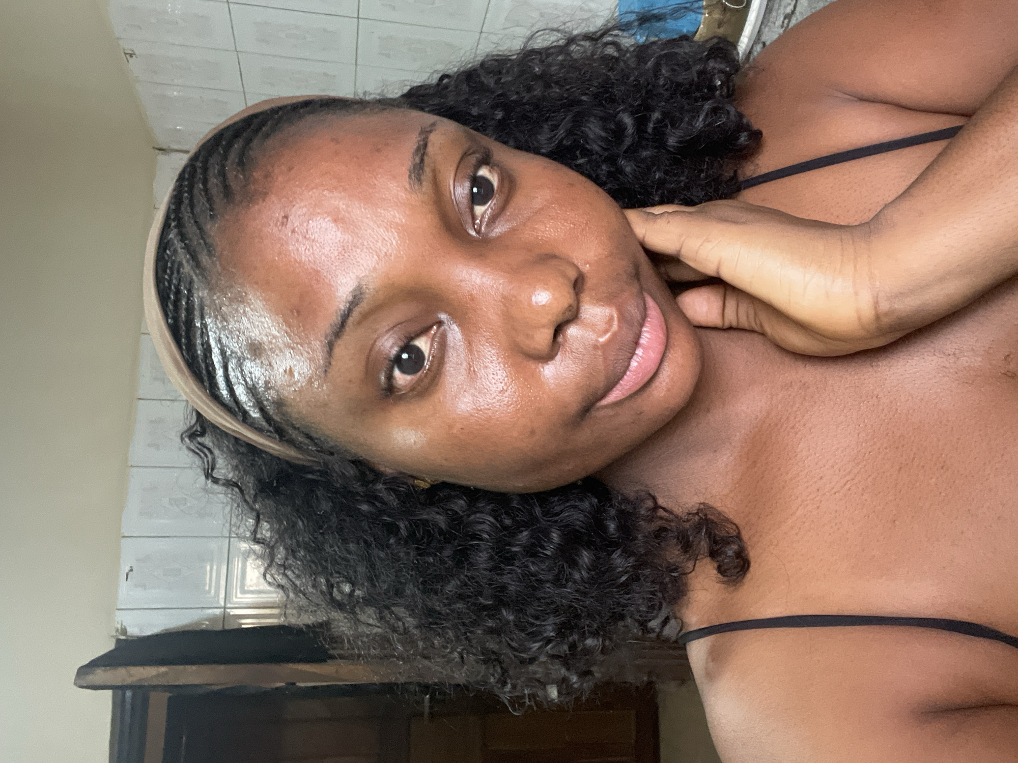 A medium close-up shows a dark-skinned woman looking at the camera. Her hair is in cornrows along the front with a brown headband and curly in the back. She is wearing a black spaghetti strap top. Her right hand is touching her neck. The location is likely indoors, potentially a bathroom, as suggested by the tiled wall. The city of Jos, Nigeria cannot be confirmed from this image.