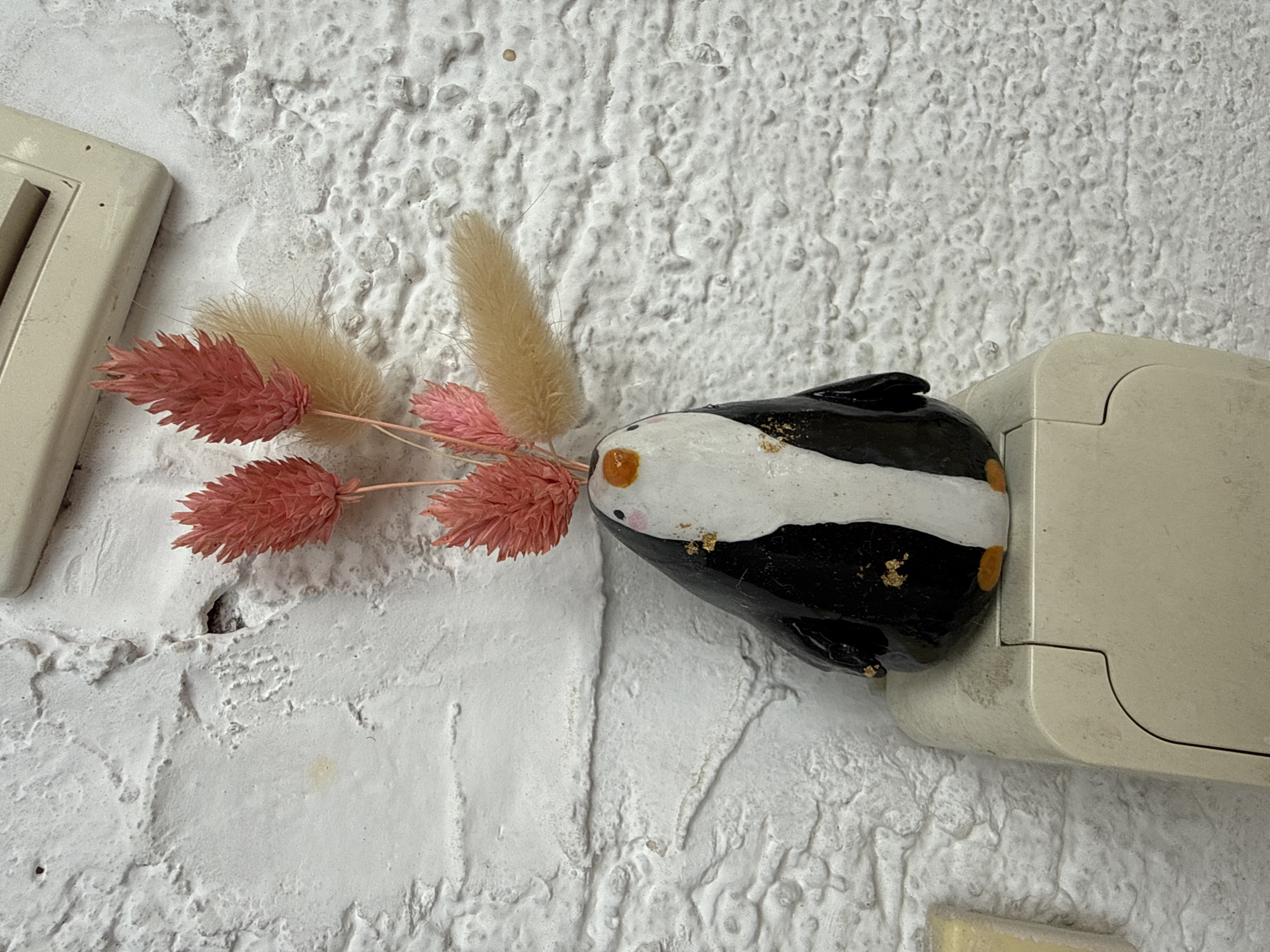 A close-up, high-angle shot shows a decorative ceramic penguin with dried pink and tan flowers. The penguin is black and white with gold accents, and its feet are orange. It is positioned on top of a beige electrical outlet. The background is a textured white wall. The scene is located in Apatin Municipality, Serbia.
