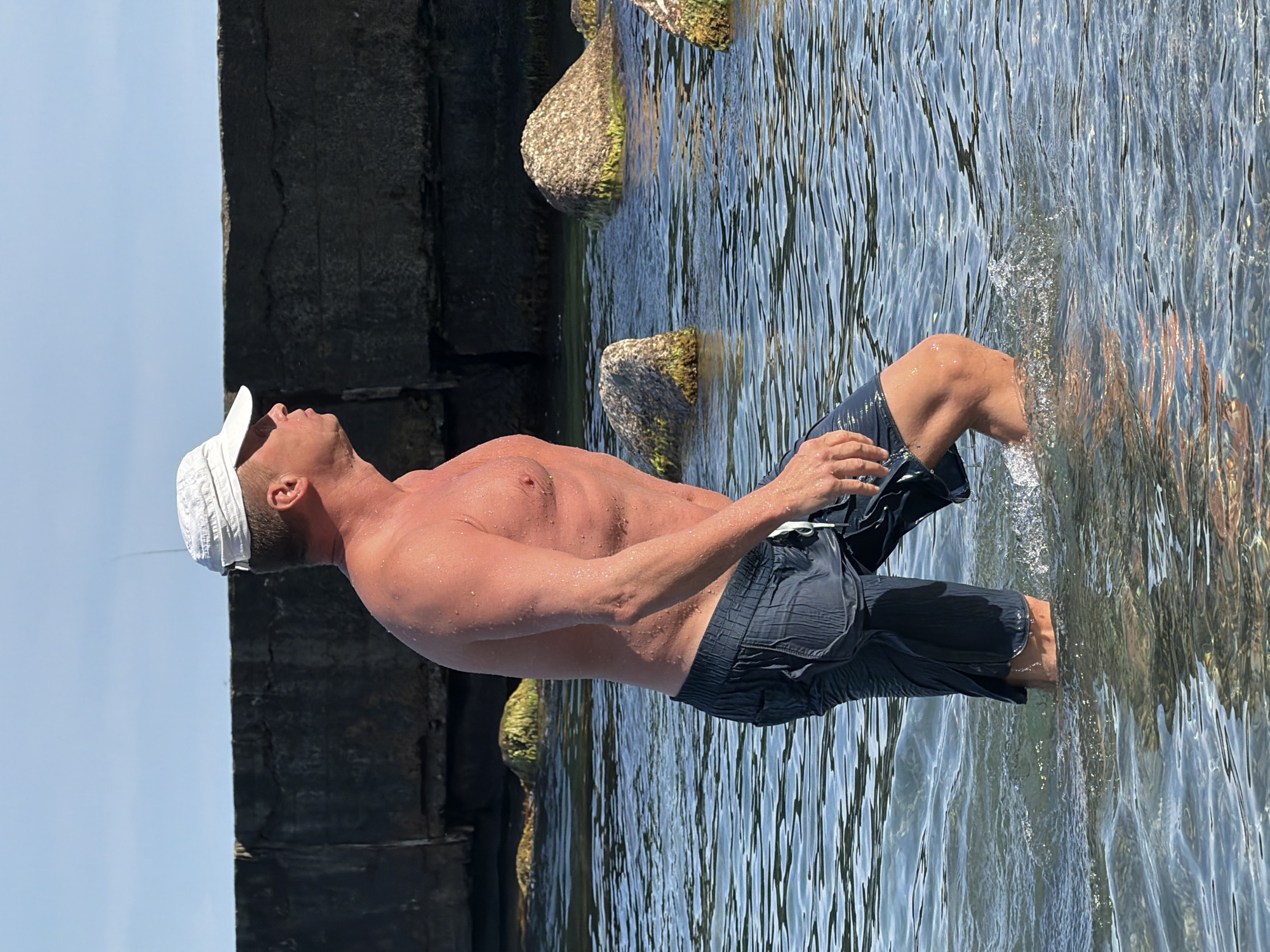 The image features a shirtless man wearing a white cap and dark shorts, standing in the shallow water near a stone structure. The man is facing upwards, with his head tilted back, suggesting he might be enjoying the sun or looking at the sky. His body is wet, with water droplets visible on his skin. The water is clear and reflects the sunlight, creating rippling patterns. Several large, moss-covered rocks are partially submerged in the water, adjacent to the stone structure which appears to be part of a breakwater or pier. The sky is a clear, bright blue. The overall scene suggests a warm, sunny day, likely during the summer. The location appears to be a coastal area, possibly a beach or a rocky shoreline. The man's posture and expression convey a sense of relaxation and enjoyment of the natural surroundings. There is no visible text in the image.
