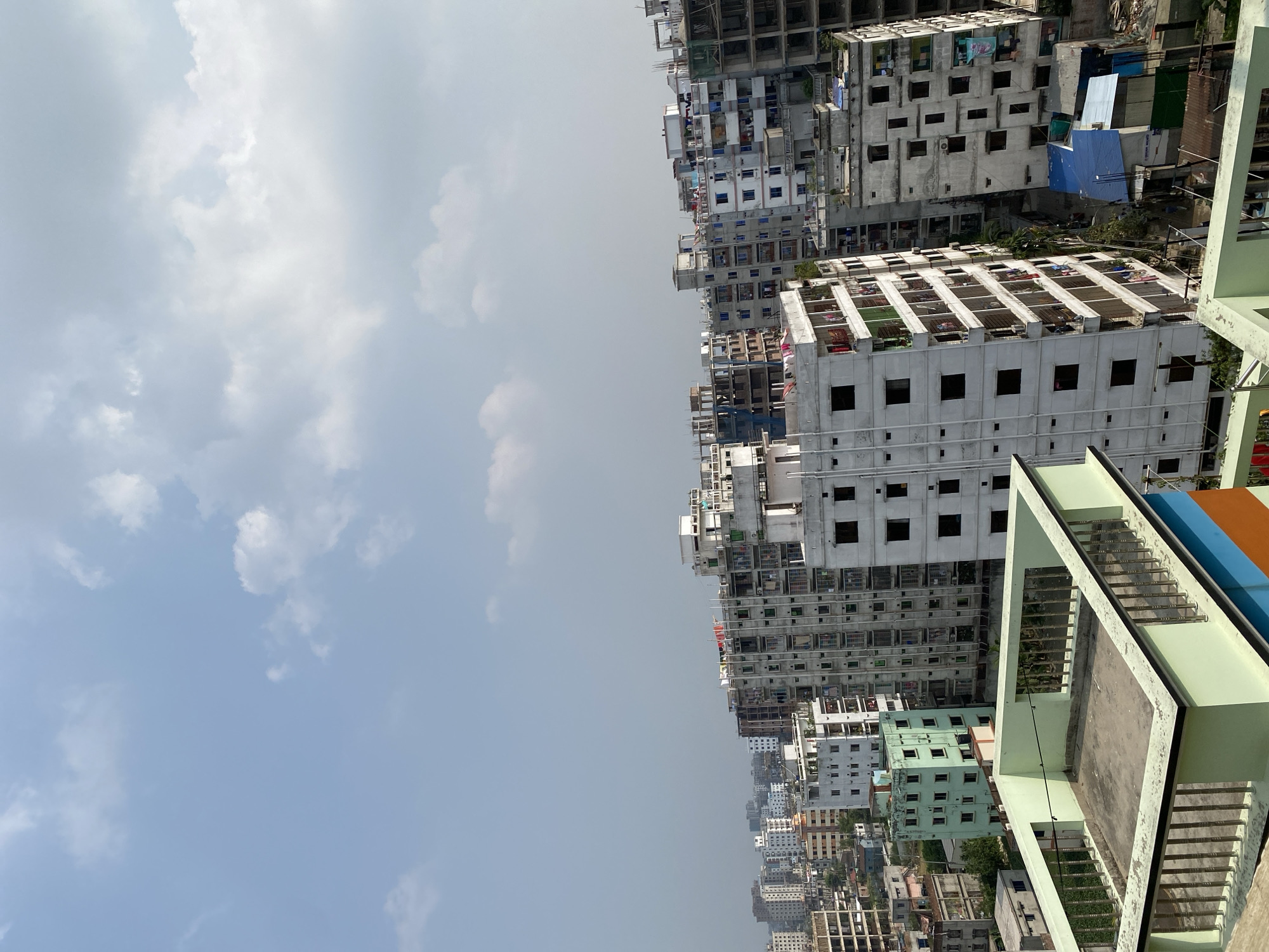 The image depicts a densely populated urban scene in Dhaka, Bangladesh, characterized by numerous multi-story buildings, many of which appear to be under construction or in varying states of completion. The buildings are predominantly concrete structures with exposed frameworks and unfinished facades. Some buildings exhibit signs of occupancy with clotheslines and personal belongings visible.
The sky is overcast with a mix of light gray and white clouds, indicating possible hazy conditions. The overall atmosphere appears humid. The perspective is taken from an elevated position, looking across the urban landscape. No people are visible.