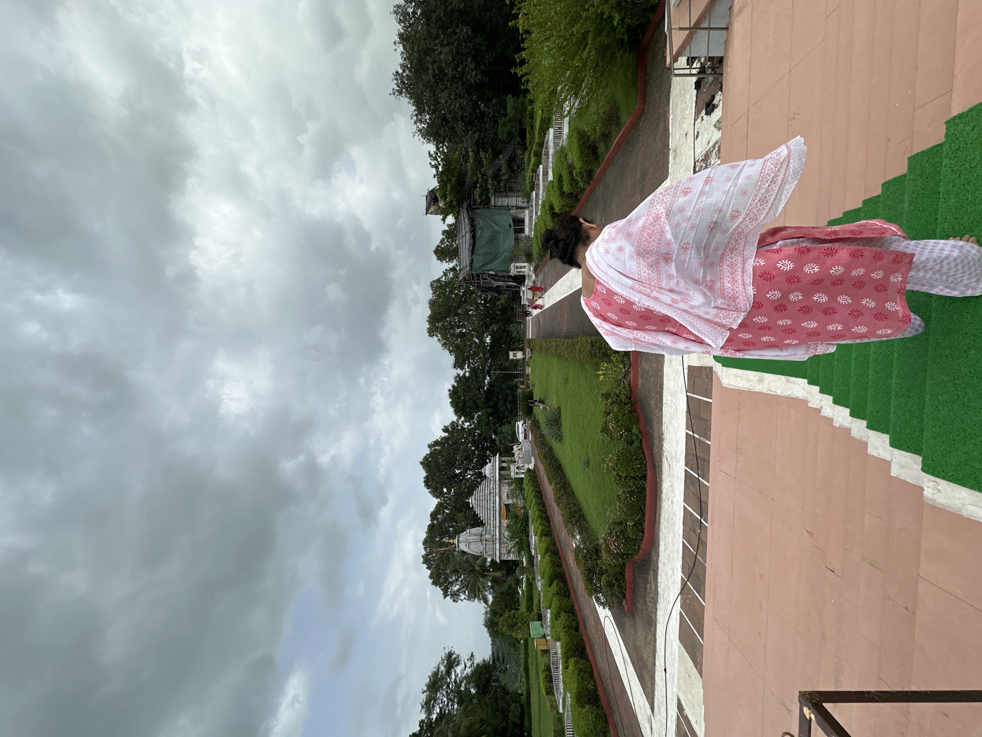A woman wearing a pink and white patterned outfit walks down a set of wide steps covered in green turf on the right and reddish-brown stone on the left. She is facing away from the camera, her back and head visible. Behind her, a large, well-maintained garden area is visible. A white temple with a pointed roof sits in the middle of the garden, surrounded by lush green grass and manicured bushes. Trees are scattered throughout the landscape. The sky is heavily overcast with dark grey clouds. The location is identified as Shahapur, India.