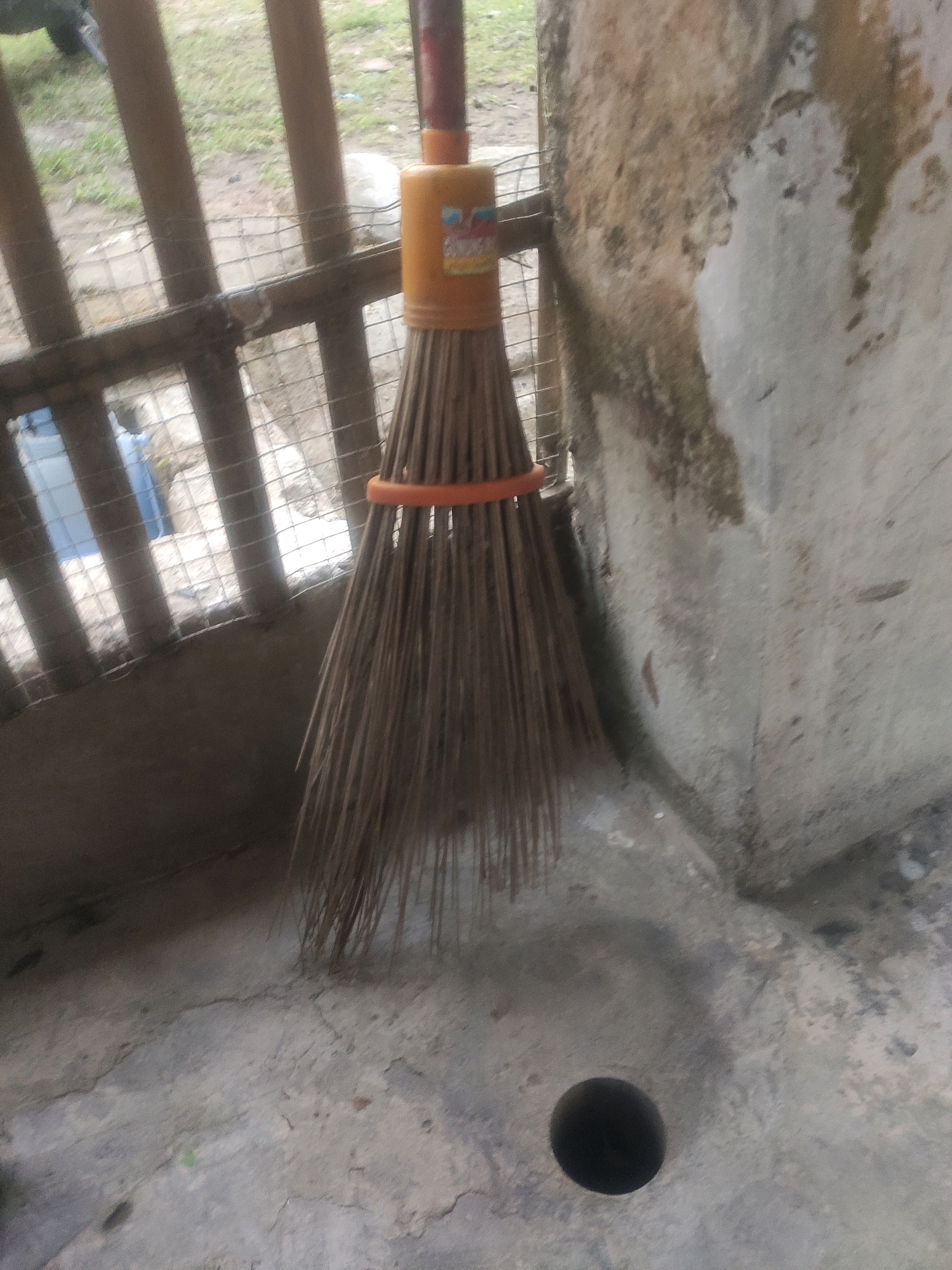 A low-angle, slightly out-of-focus medium shot shows a broom with a plastic orange handle leaning against a weathered concrete wall. The broom bristles are made of natural material, likely coconut fronds, and are bound together with an orange plastic ring. The broom leans against a portion of the wall that exhibits a mottled texture with patches of light and dark coloration, possibly due to moisture or dirt. To the left of the broom, a rustic fence made of vertical wooden posts and horizontal wooden slats can be seen. Behind the fence, a glimpse of an outdoor environment with sparse green vegetation is visible, along with what appears to be a blue plastic container. The ground in the foreground is concrete, cracked and uneven, with a dark, circular opening in the lower right quadrant of the image, resembling a drain or a hole in the ground. The lighting is somewhat dim and diffused, suggesting an overcast day or shaded area. The scene appears to be an outdoor, utilitarian area, possibly a backyard or alleyway, in a tropical or subtropical climate.