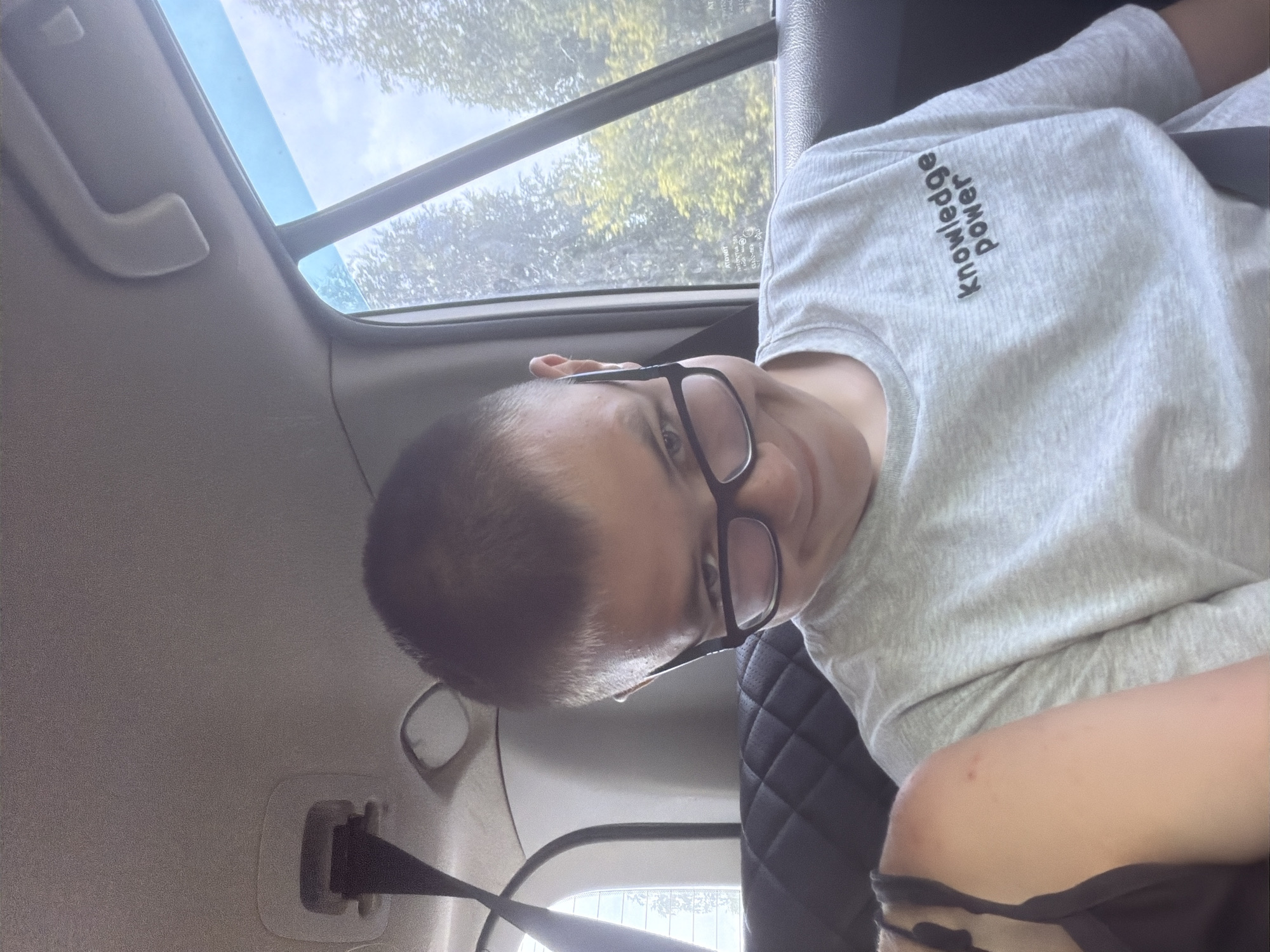 A young person wearing glasses and a grey t-shirt with the text "knowledge power" on it is sitting in the back seat of a car. The person is looking towards the camera with a slight smile and head tilt. The car's interior is visible, including a black quilted seat and a seatbelt. Through the window, trees and a cloudy sky are visible, suggesting an outdoor setting, likely in Nadezhdino, Russia. Another person's shoulder is visible on the left side of the frame.