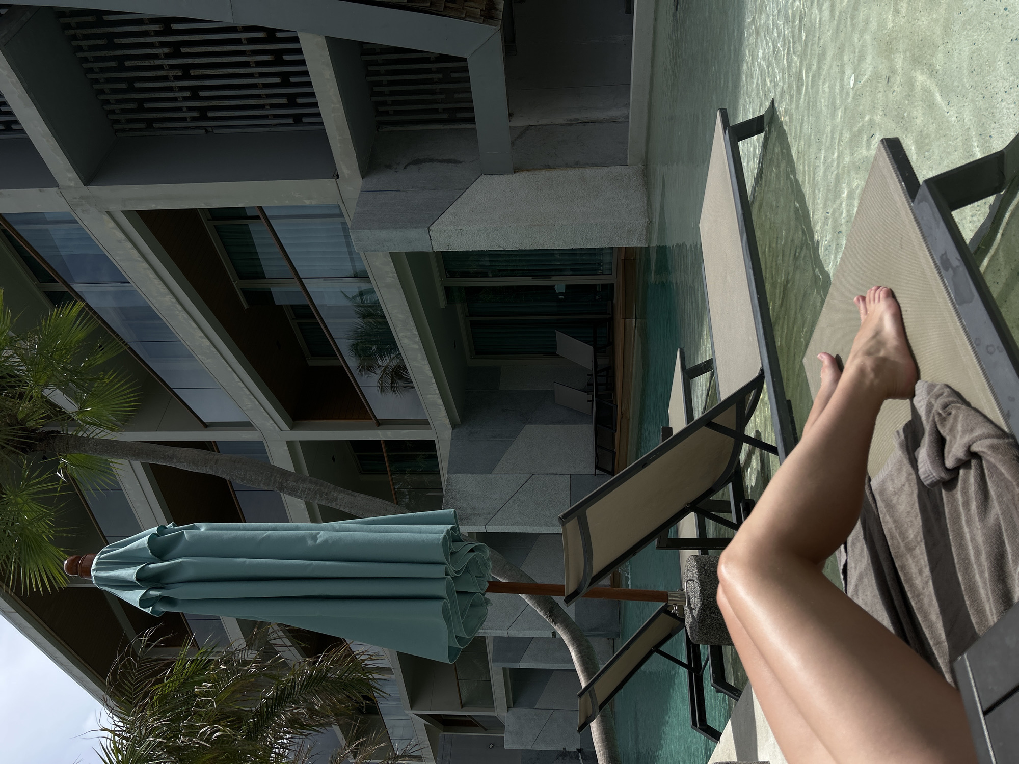 The scene shows the legs of a person reclining on a chaise lounge adjacent to a swimming pool, likely at a resort or hotel. The location appears to be in Rawai, Thailand, based on the architectural style of the surrounding buildings and the presence of palm trees. The person's legs are angled towards the viewer, and they are partially covered by a towel. The chaise lounge is dark-colored, and there is an umbrella providing shade. The building in the background is multi-story, with a modern design featuring concrete and glass. Palm trees are visible beside the building, suggesting a tropical environment. The pool water appears clear and reflects sunlight.