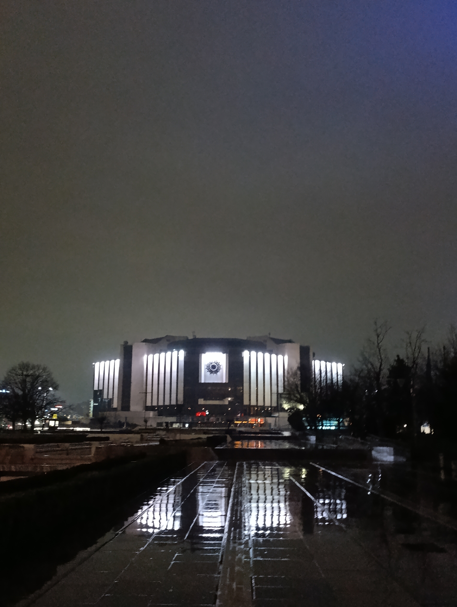 The image shows the National Palace of Culture in Sofia, Bulgaria, at night. The building is illuminated with vertical white lights. The reflection of the building is visible in a water feature in the foreground. The sky is overcast and dark. Trees are visible on the right side of the image, silhouetted against the night sky.