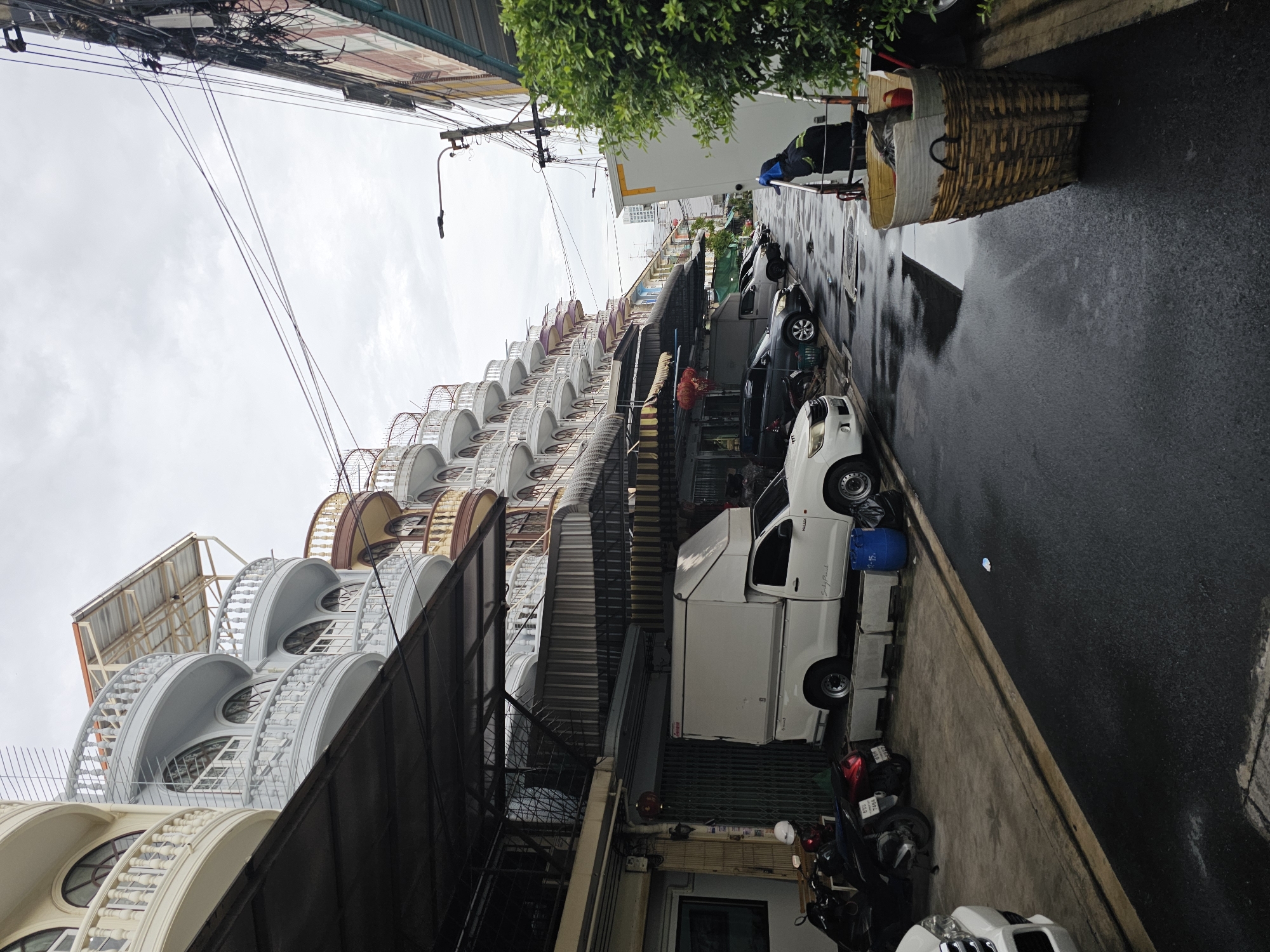 The image depicts a narrow street in Bangkok, Thailand, likely a residential or commercial area. The overcast sky suggests it might be raining or has recently rained, as the asphalt road is wet and reflective, with puddles present.
On the left side of the street, there are multi-story buildings with distinctive architecture, featuring rounded balconies with ornate railings and arched windows. These buildings appear to be a row of shops or apartments. Below the upper floors, there are awnings and covered areas, indicating ground-level businesses. Several motorcycles are parked along the sidewalk.
A white pickup truck with a white camper shell is parked in the street. Further down the street, other vehicles, including cars and motorcycles, are visible, also parked.
On the right side of the street, more buildings are visible, including what appears to be a store or restaurant with a green awning and lush vegetation. A large woven basket, possibly used for collecting items, is placed near the curb. A person wearing dark clothing and blue gloves is standing next to a white truck. Electrical wires are strung across the street, a common sight in Bangkok.
The overall scene is one of everyday urban life in Bangkok, characterized by dense architecture, mixed-use buildings, and the presence of vehicles and pedestrians.