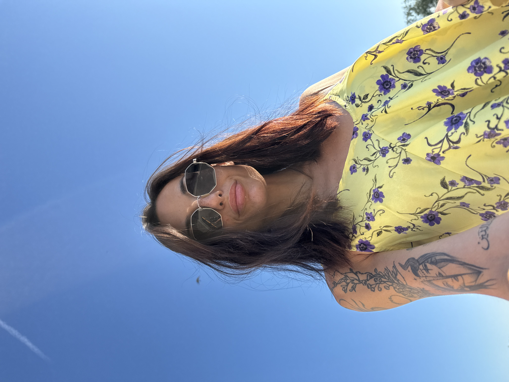 A woman with long, dark hair and sunglasses is captured in a low-angle, selfie-style shot against a clear blue sky. She is wearing a yellow sundress with a purple floral pattern. Her left arm, adorned with tattoos, is visible. The image does not provide any specific landmarks or details to place it within the city of Pleven, Bulgaria.