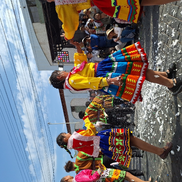 A vibrant cultural celebration unfolds with a group of people, mostly young women, adorned in traditional outfits. They wear colorful skirts, yellow blouses, and patterned tops. Some are throwing white confetti into the air. Spectators are seen in the background, with some wearing hats. The scene is illuminated by stage lights, set against a backdrop of a blue sky, power lines, and a building.