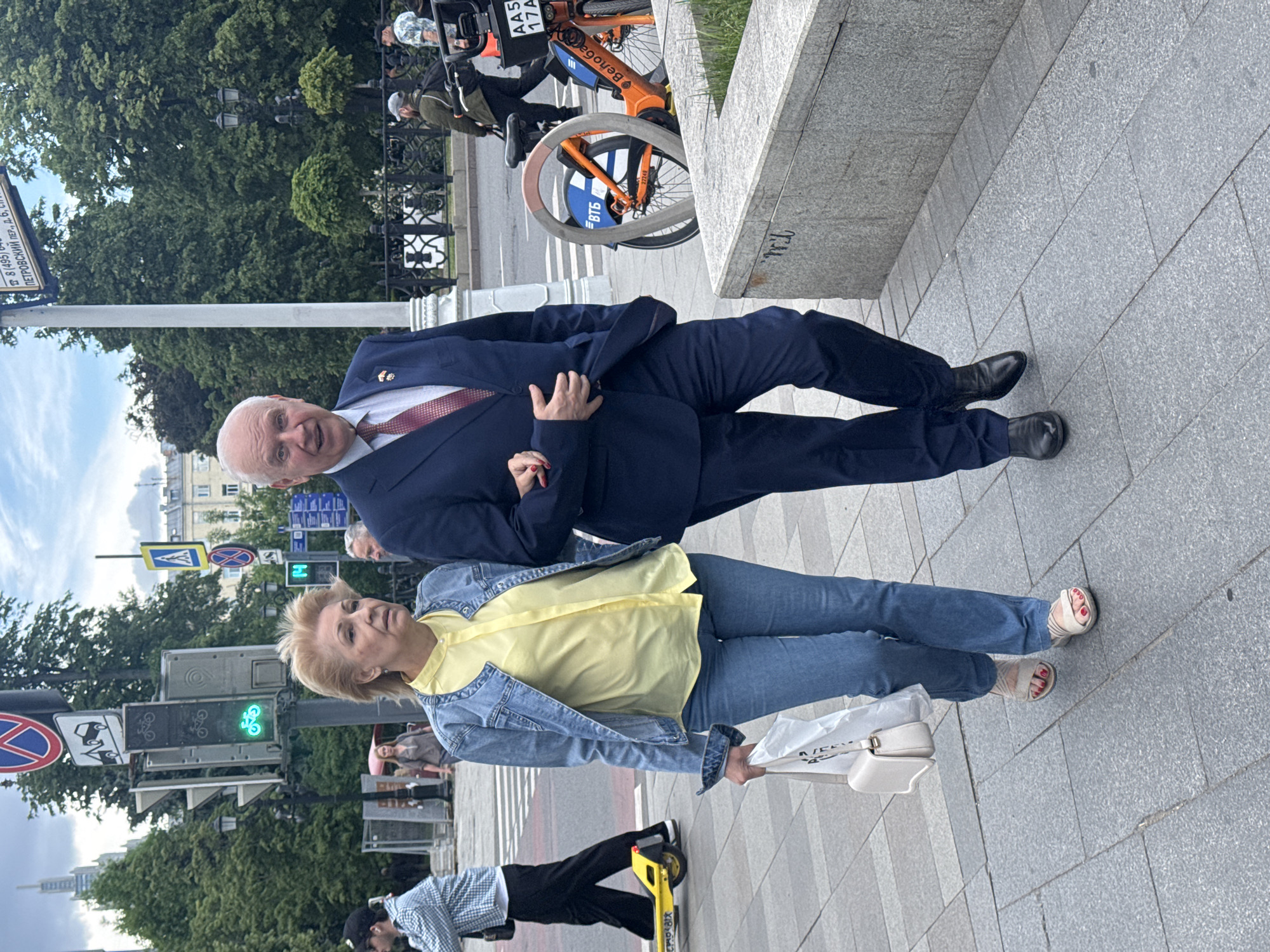The image shows a man and woman standing on a city sidewalk. The man wears a navy blue suit with a red tie and black shoes. The woman wears a yellow shirt, a denim jacket, jeans, and light-colored sandals. She holds a white bag.
In the background, there's a street sign indicating "Петровский пер., д. 6," which translates to Petrovsky Lane, building 6, placing the scene in Moscow, Russia. There is a bicycle stand, a street light with a green bicycle signal, and other pedestrians.