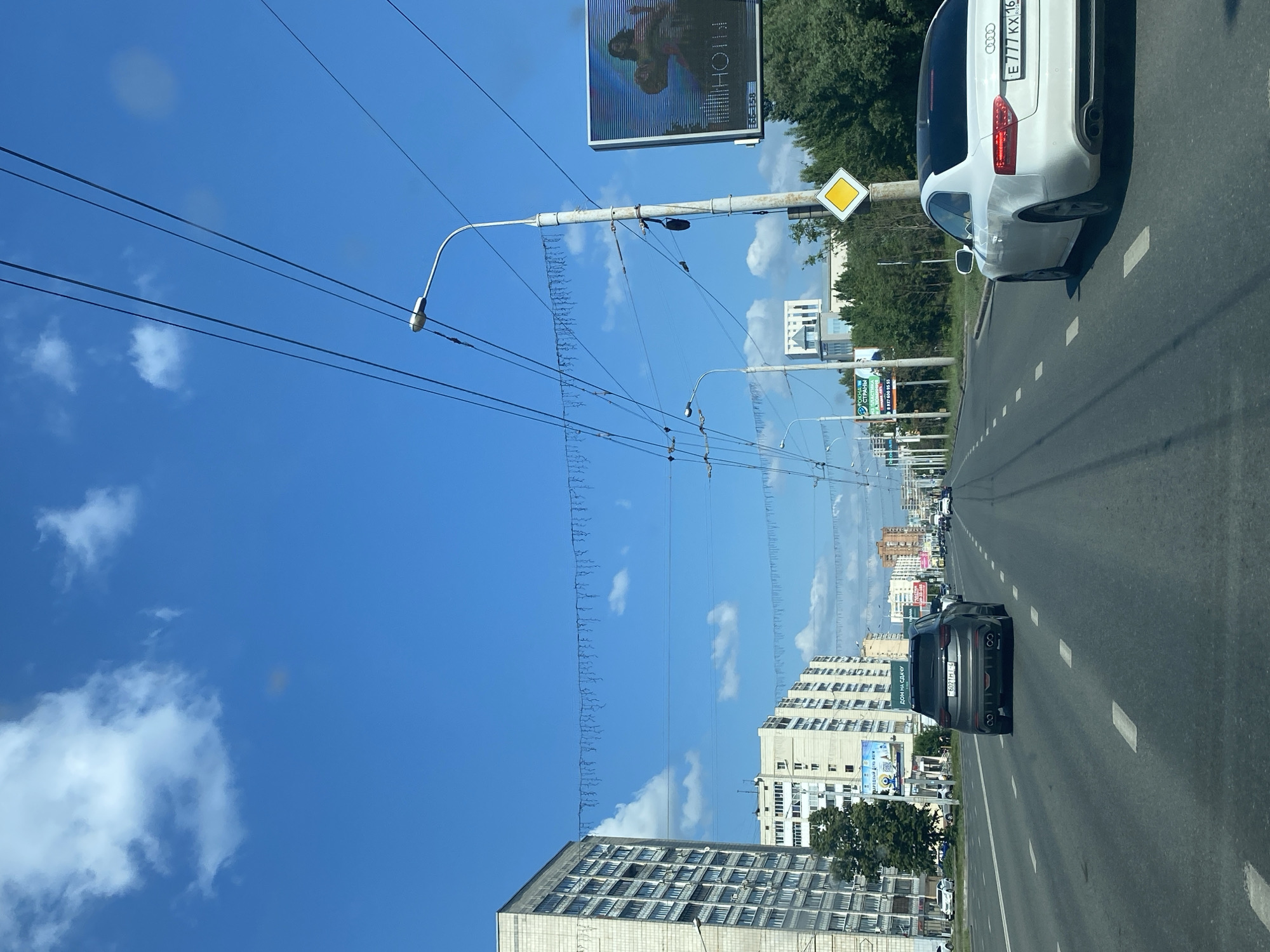 The image shows a street view in Kazan, Russia, on a sunny day. The perspective is from inside a moving vehicle, looking forward. On the left, a tall, light-colored apartment building with many windows lines the street. Ahead, several similar apartment buildings are visible, interspersed with trees and streetlights. The road has multiple lanes marked with white dashed lines.
In the foreground, a dark-colored SUV is traveling in the same direction as the viewer. Further ahead on the right, a white Audi coupe is also in motion. Overhead, a network of power lines crisscrosses the sky, supported by poles. A large digital billboard displaying an advertisement is visible on the right side of the road. The sky is blue with scattered white clouds.