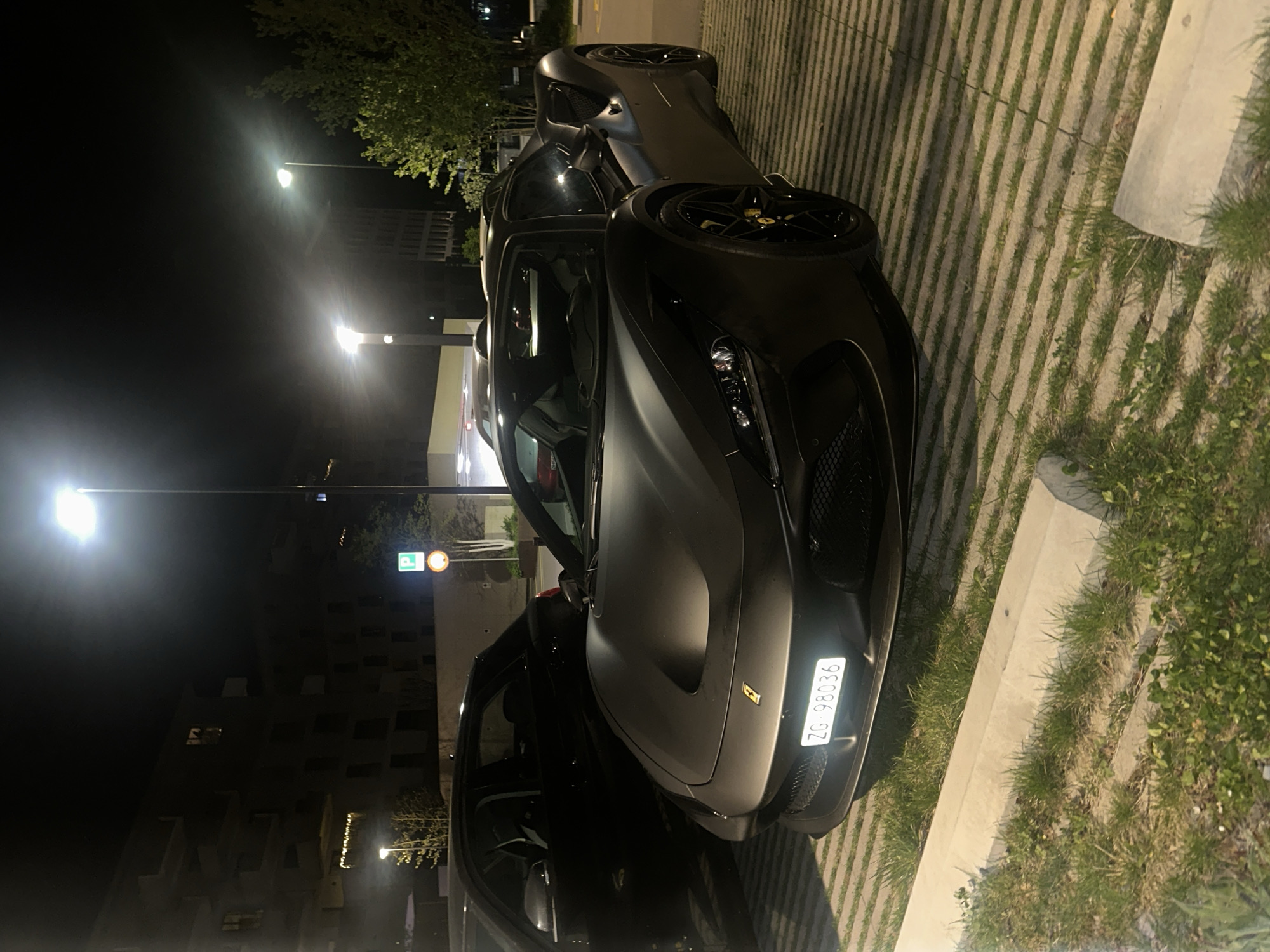The image captures a nighttime outdoor scene, likely in a parking area in Dübendorf, Switzerland, as suggested by the Swiss license plate format. Two luxury cars are parked side-by-side under the illumination of streetlights.
The primary subject is a sleek, matte dark grey or black Ferrari positioned in the foreground, facing slightly towards the viewer. Its aerodynamic lines, intricate front grille, and modern headlights are visible. The car features large dark alloy wheels with distinctive yellow brake calipers and a small yellow Ferrari emblem on the front hood. A clear white license plate, "ZG 98036", is prominently displayed on the front bumper. "ZG" is the canton code for Zug, Switzerland.
Behind and slightly to the left of the Ferrari, another dark-colored vehicle, possibly a luxury SUV or sports car, is parked. Fewer details are discernible for this second vehicle due to its position and the shadows.
The parking surface consists of permeable pavers with strips of grass growing between the concrete grids, indicating an environmentally conscious design. A concrete curb runs along the edge of the parking area, bordered by more grass and some low-lying foliage. The scene is well-lit by several bright streetlights, which cast a luminous glow and create defined shadows. In the background, multiple-story buildings, possibly residential or commercial, are visible, with some windows illuminated. A blue circular parking sign with a white 'P' is also noticeable in the mid-ground. The night appears clear, with no adverse weather conditions visible.