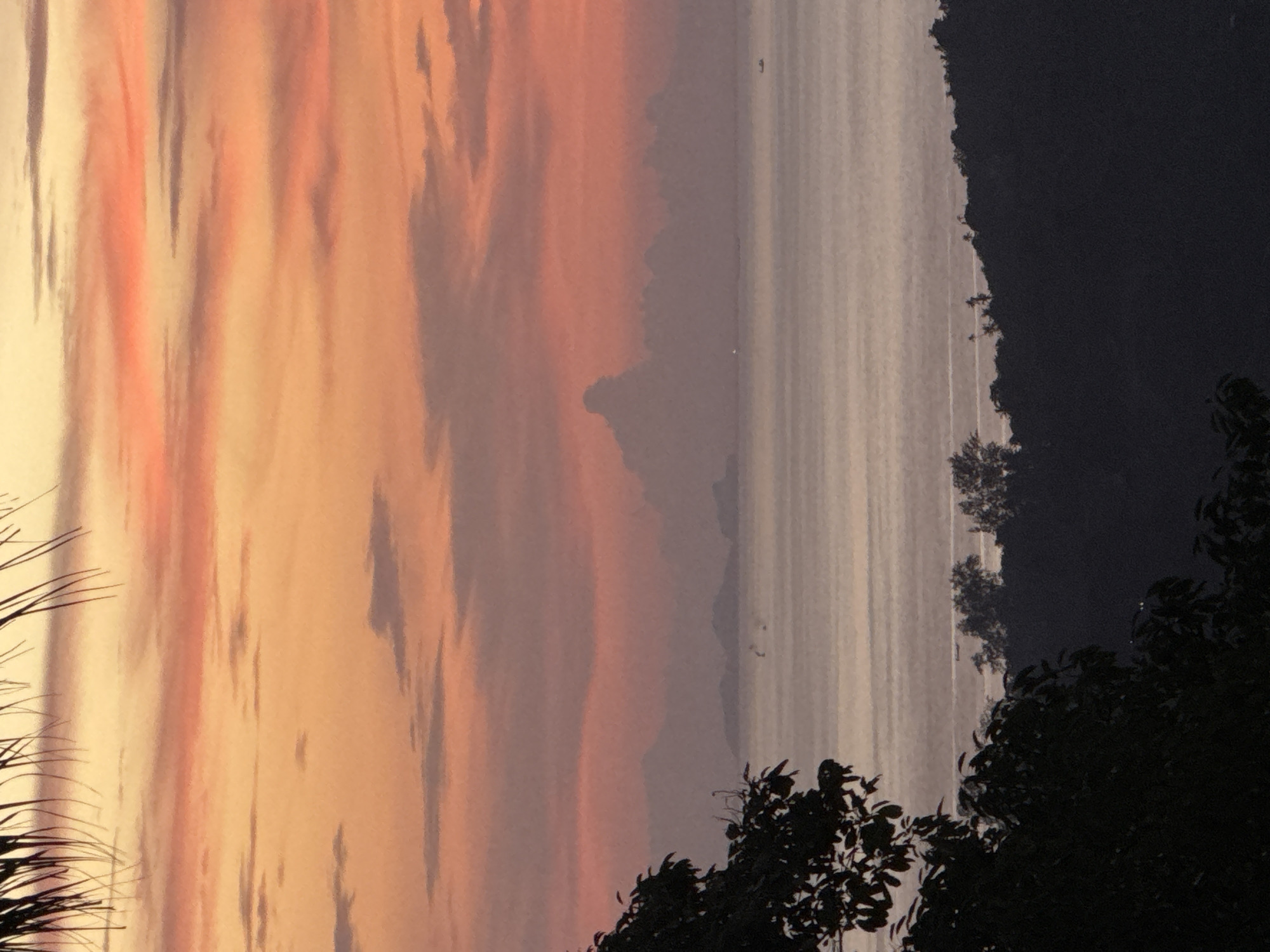 The image depicts a serene sunset scene over the ocean, likely viewed from an elevated position on Ko Pha-ngan District, Thailand. In the foreground, the dark silhouette of lush vegetation, possibly a tree with leaves and branches, frames the left side of the composition. Beyond this, a broad expanse of calm ocean stretches towards the horizon, its surface reflecting the muted colors of the sky. Scattered across the water, faint silhouettes of small boats or islands can be observed. In the distance, a hazy silhouette of landforms, possibly more islands or coastal mountains, emerges from the ocean. The sky is a canvas of warm hues, transitioning from soft orange and pink near the horizon to a pale yellow at the top, with wisps of clouds adding texture and depth to the sunset. The overall atmosphere is tranquil and picturesque.