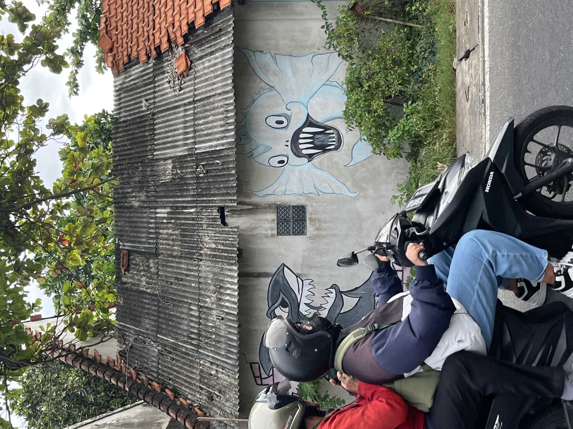 In Kuta Selatan, Indonesia, two individuals ride a black Honda motorcycle. The person in the foreground, driving the motorcycle, wears a black helmet, a dark jacket with white and blue accents, and blue jeans. They are also wearing black and white sneakers. The passenger behind them wears a lighter colored helmet and a red jacket. The motorcycle is on a paved road, next to a sidewalk with overgrown grass and a concrete barrier. Behind the road and sidewalk is a wall with two murals. The mural on the left depicts a shark with sharp teeth, and the mural on the right shows a light blue, cartoonish fish with large eyes and sharp teeth, reminiscent of a character from an anime or cartoon. The wall is partially covered by a corrugated metal roof. Green trees and branches overhang the scene from above, with some leaves casting shadows. The sky is overcast.