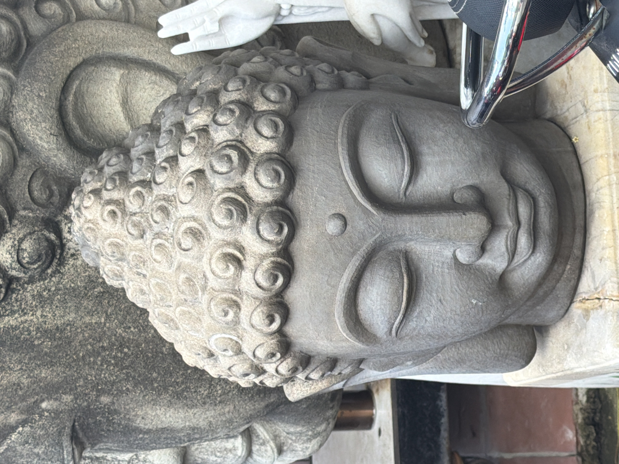 The image shows a close-up of a stone Buddha head. The Buddha head is in the foreground, with its eyes closed and a serene expression. The top of the head is covered in curly hair. To the right of the Buddha head, a white stone hand is visible, with its palm facing forward. In the background, another large stone structure, possibly another Buddha statue, is partially visible. The Buddha head is placed on a stone pedestal. A shiny metallic object, likely part of a motorcycle or similar item, is in the lower right corner of the image. The lighting suggests an outdoor setting. Without further context or identifying landmarks, the specific location in Da Nang, Vietnam, cannot be determined from the image alone.