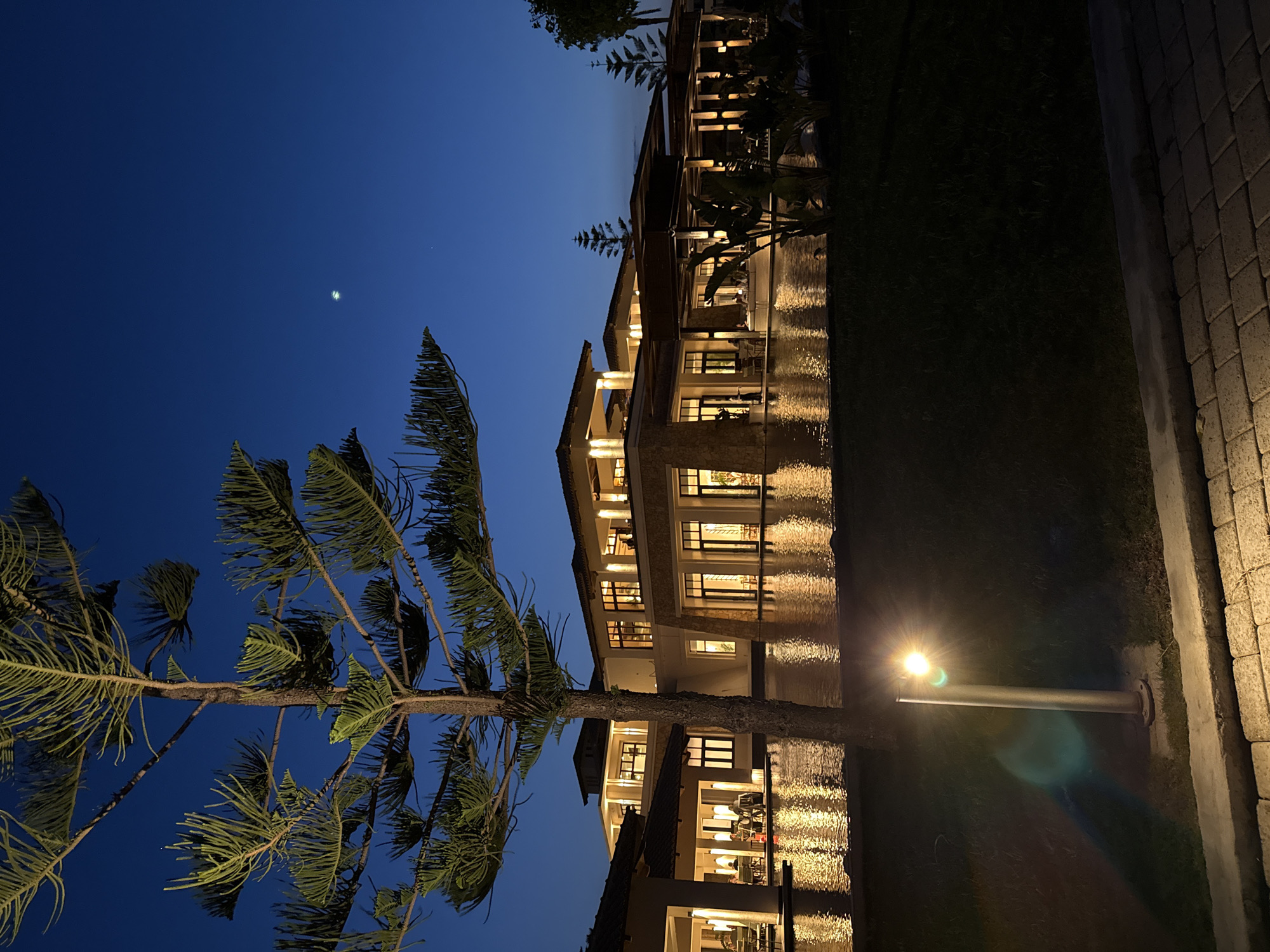 This is an outdoor nighttime shot showing a multi-story building in Kos, Greece. The building appears to be a hotel or resort with multiple illuminated windows and balconies. The image is taken from ground level, with a tree in the foreground and the building extending into the background. The sky is dark blue, suggesting twilight or early evening, and there is a bright object in the sky, likely the moon. The lights from the building reflect in the water below, creating a symmetrical effect. The architecture of the building features arches, stone detailing, and multiple levels. A streetlamp is located in the lower right corner of the frame. There are people in the building as evidenced by their silhouettes visible inside.