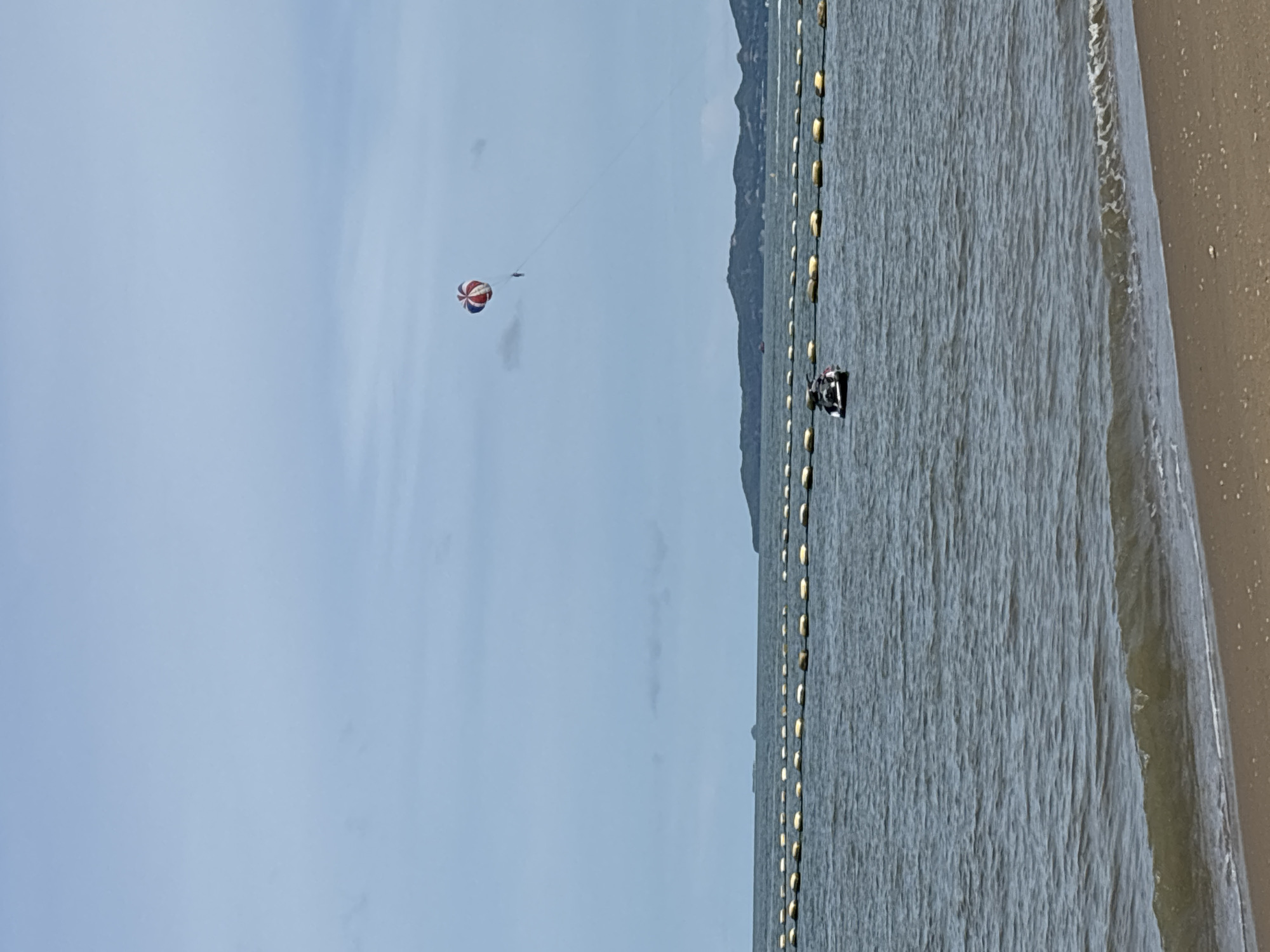 The image depicts a coastal scene in Pattaya City, Thailand. In the upper portion of the frame, a parasail, featuring red and white segments, is visible against a pale blue, overcast sky. Below the parasail, a person is suspended. In the middle ground, a jet ski is in operation on the water, with its rider visible. A line of yellow buoys stretches across the water, separating areas of the sea. In the background, a hilly, green coastline under the hazy sky suggests the mainland. The foreground shows a sandy beach with small waves gently lapping the shore. The overall atmosphere is calm and typical of a beach activity day.