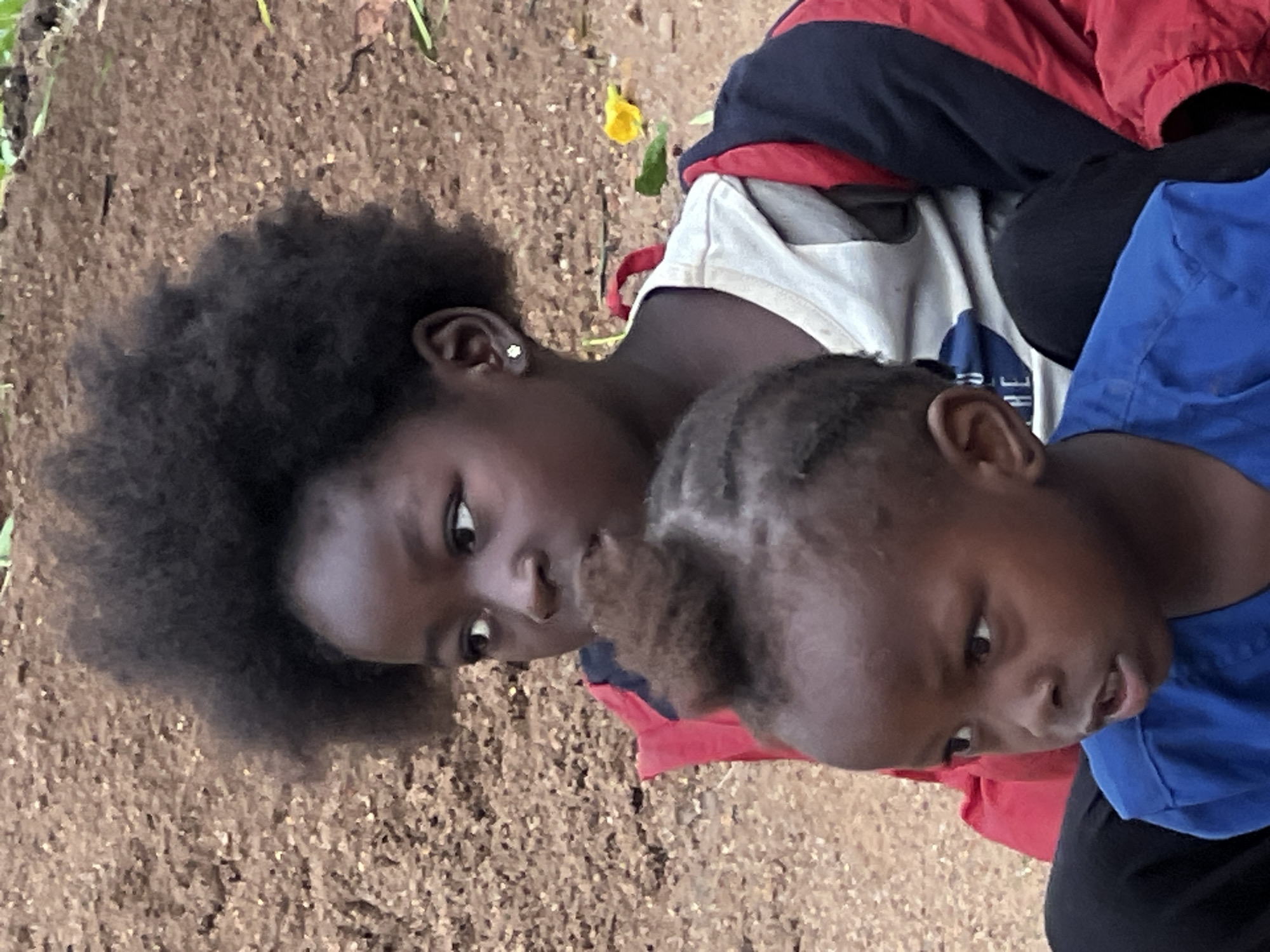 The image shows two young children, likely siblings, sitting outdoors. The older child, in the background, has dark, voluminous curly hair and is wearing a red and blue jacket over a white t-shirt. They are looking to the left with a thoughtful expression. A small, stud earring is visible in their left ear. The younger child, in the foreground, has short, dark hair styled with a small tuft on top. They are wearing a bright blue t-shirt and are looking to the right with their mouth slightly open, as if speaking or reacting to something. The background consists of textured brown earth, with some scattered green foliage and a yellow flower visible. The scene suggests a casual, outdoor setting in a natural environment. The provided information indicates this photograph was taken in the city of Jos, Nigeria.