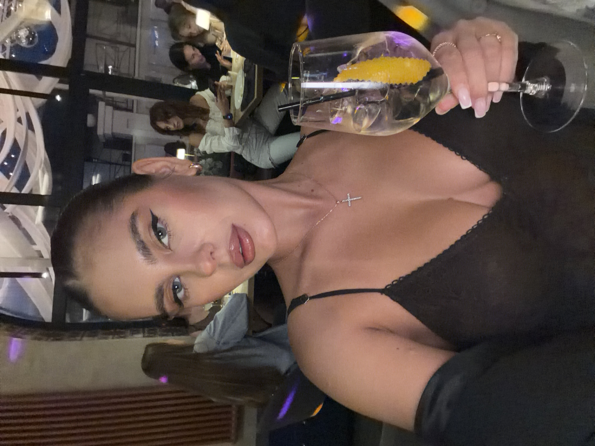 A medium shot captures a woman holding a drink with a lemon peel and a straw. She is wearing a black lace top and a necklace with a cross pendant. In the background, other people are seated at tables in a dimly lit establishment, possibly a restaurant or bar. The scene is set in Krasnoyarsk, Russia.